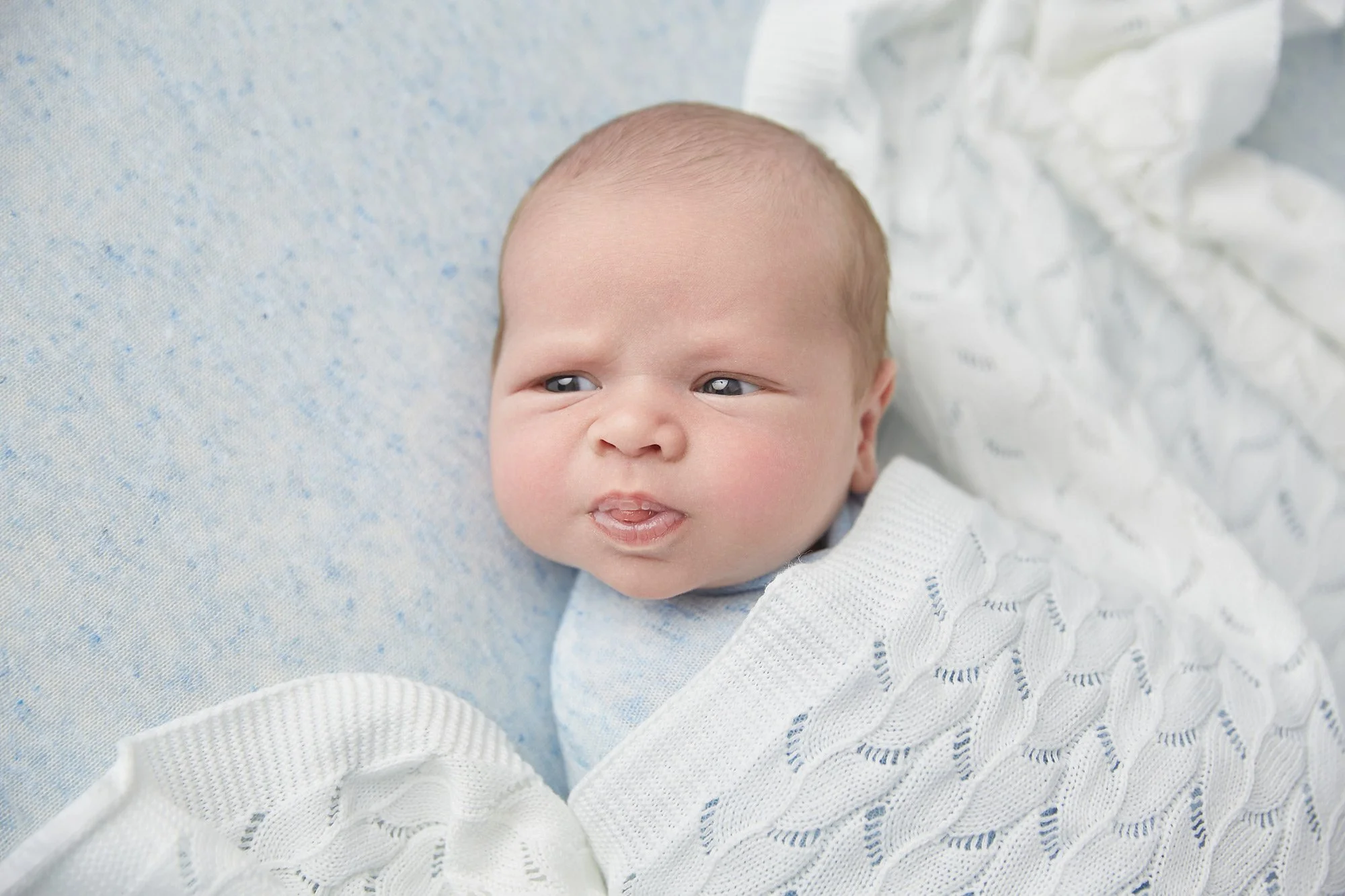 Close-up of a newborn baby with light skin and blue eyes, lying on a soft surface, wrapped in a white knitted blanket, making a curious facial expression.