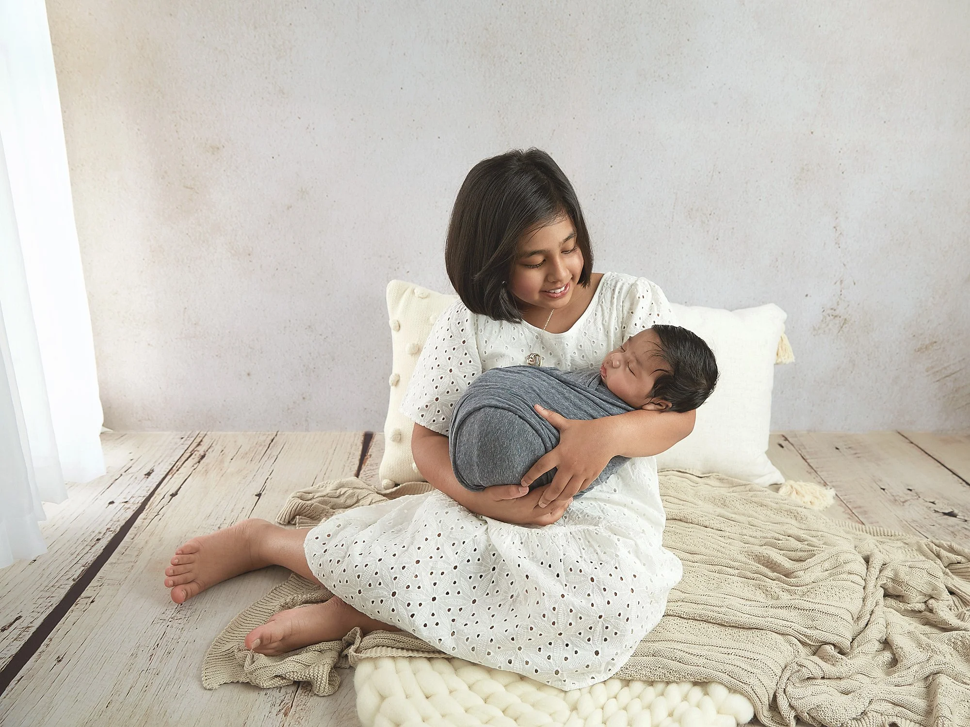 A young girl in a white dress sitting on beige blankets, holding a newborn baby wrapped in a gray blanket, in a cozy room with a white wall and pillows.