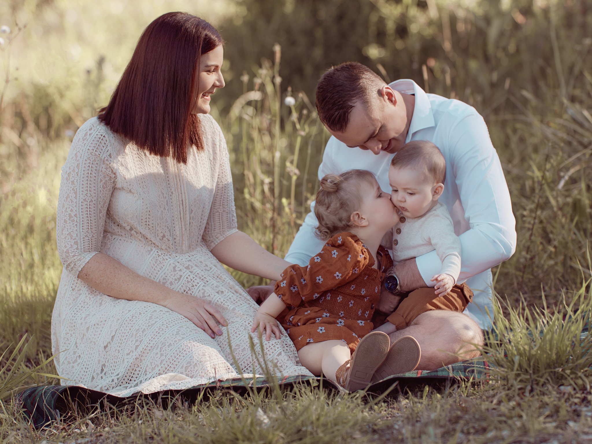 Family of four sitting on a blanket in a grassy outdoor setting; mother with brown hair in a white dress, father with dark hair in a white shirt, daughter and son with light hair, playing and sharing a kiss.
