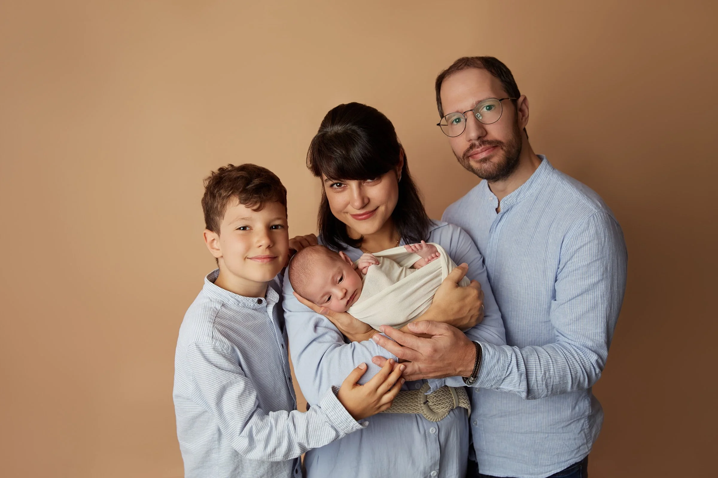 Family of four holding newborn baby, smiling, standing against a beige background.