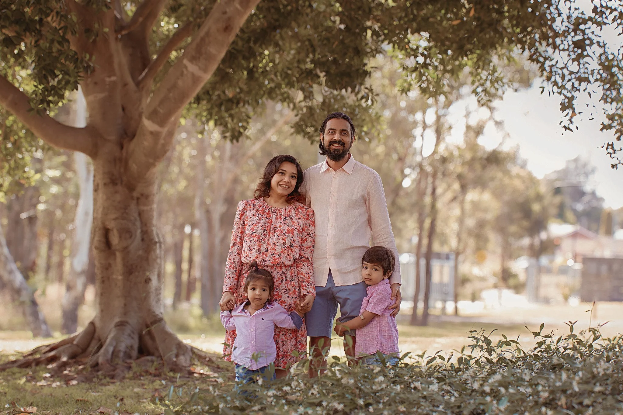 Family of four standing outdoors near a large tree, smiling at the camera in a park.