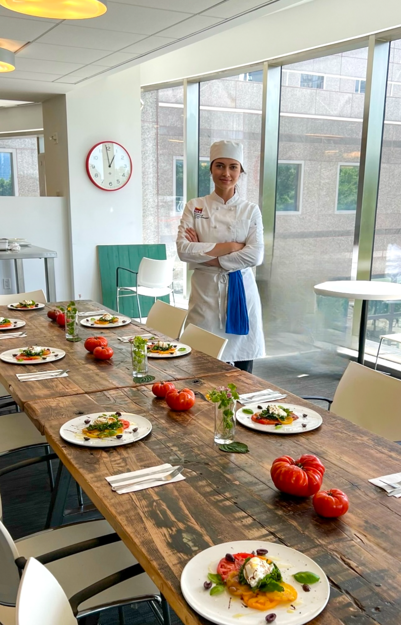 A female chef in a white uniform and hat standing with arms crossed in a modern, bright restaurant or kitchen. The long wooden table is set with plates of colorful salads, and decorated with small tomatoes and potted plants. Large windows let in natural light facing a cityscape.
