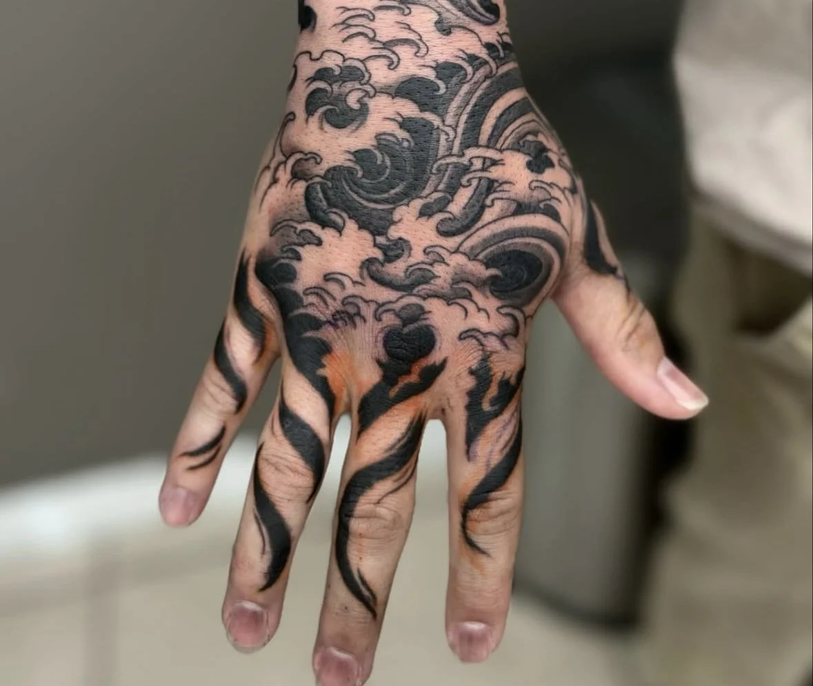 A hand with a black ink tattoo of swirling clouds and stylized flames stretching from the wrist to the fingers.