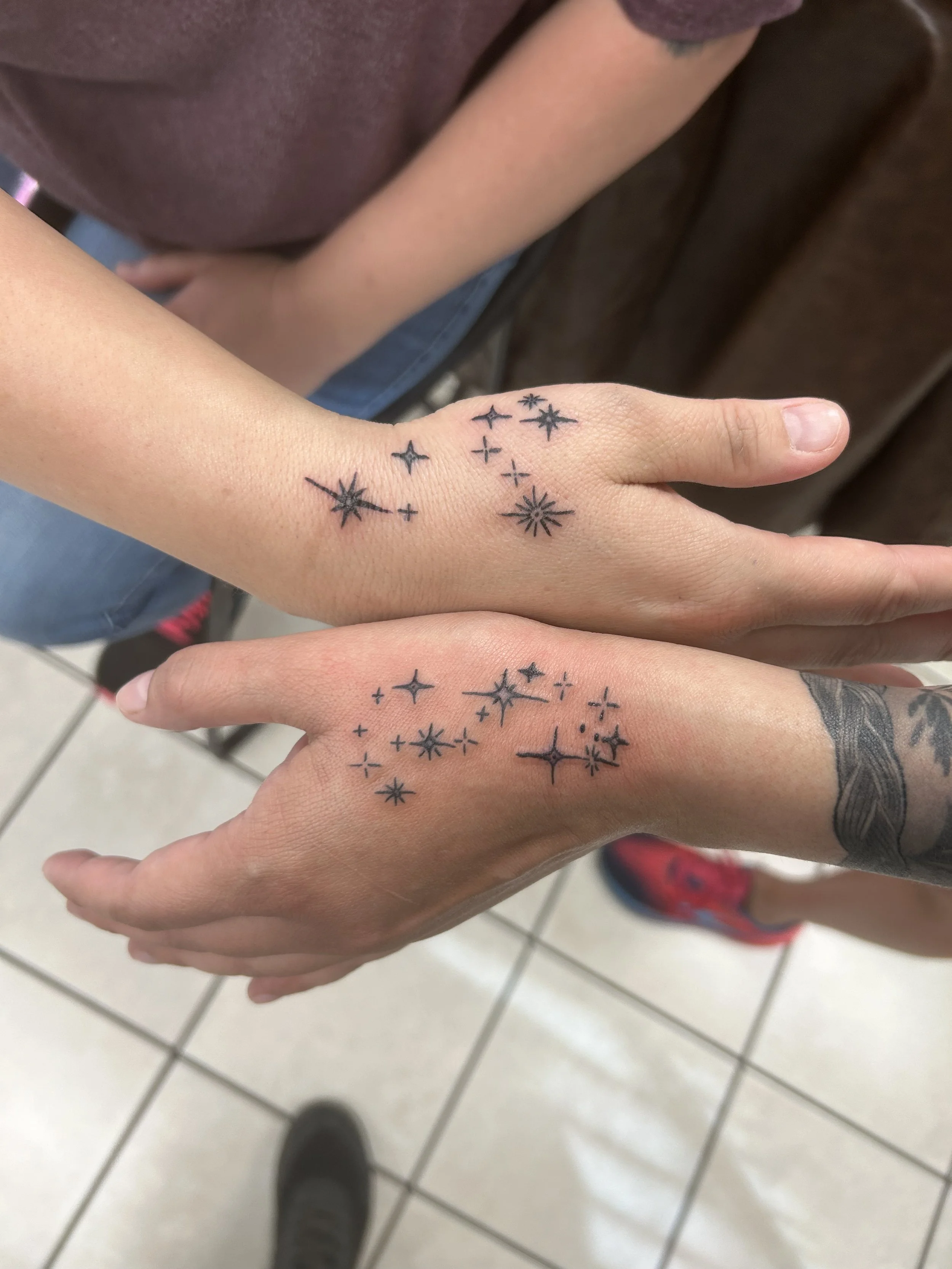 Two hands with matching black star and sparkle tattoos on the wrists, overlapping each other. One hand has a tattoo of an arm with a sleeve.