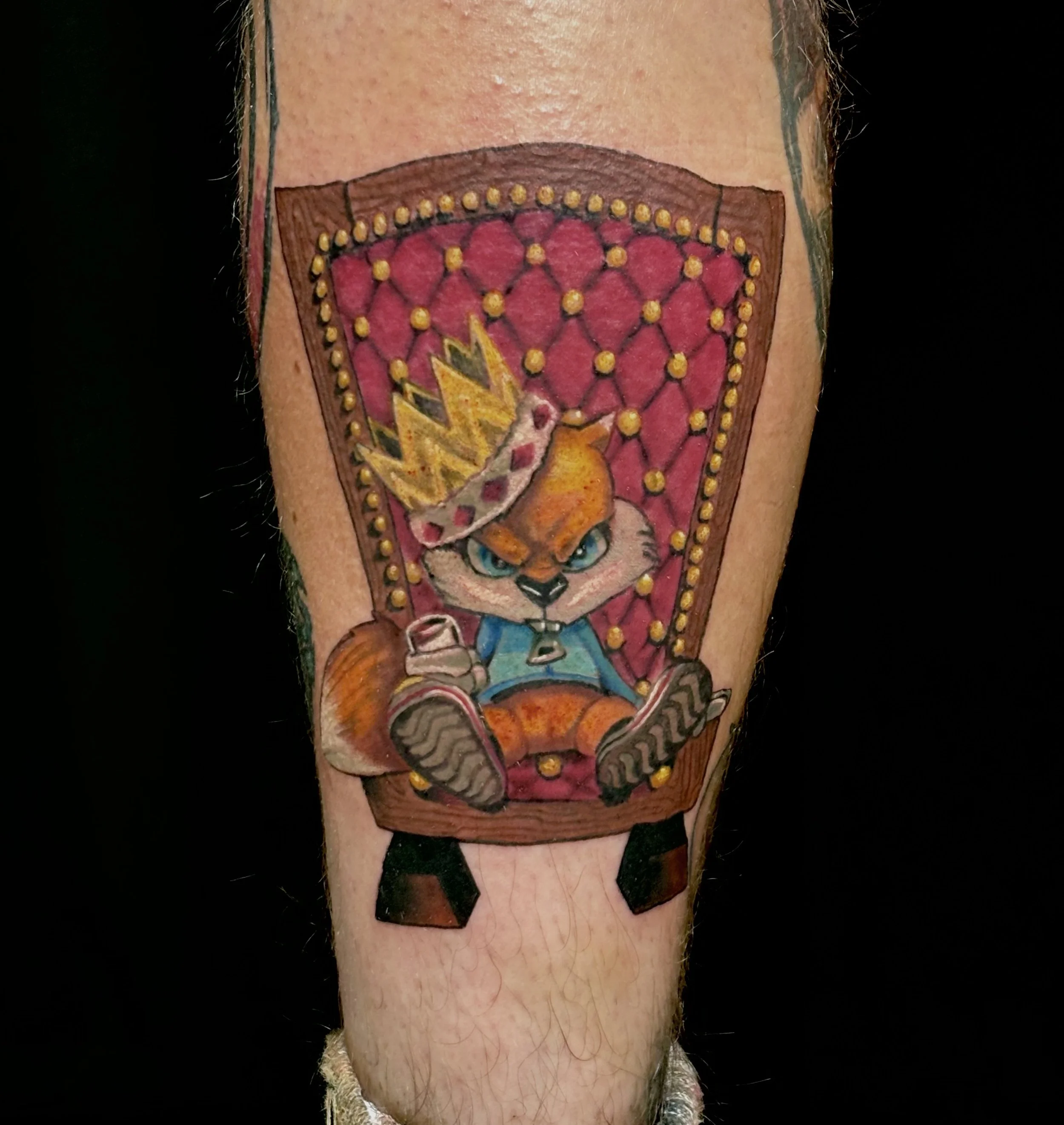 Tattoo of a cartoon tiger with blue eyes, sitting on a red throne, wearing a crown, with a blue shirt and gray shorts, on a person's arm.
