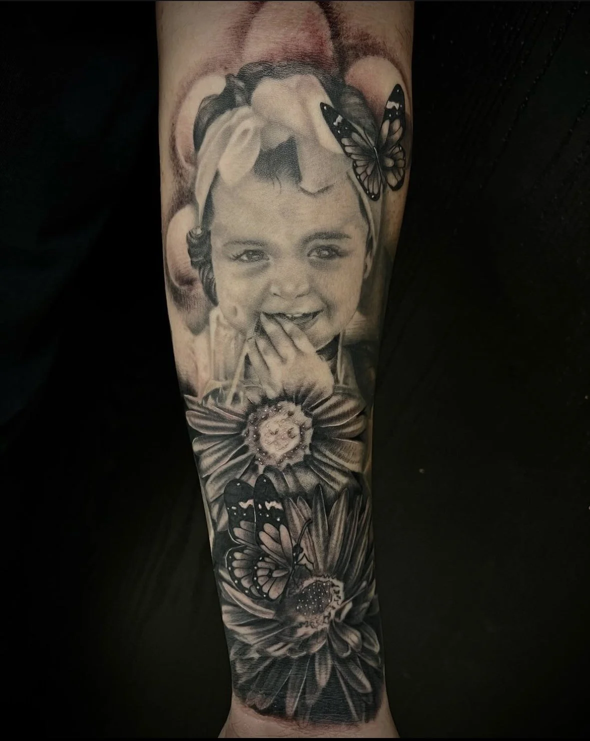 Black and gray tattoo of a smiling young girl with butterflies and flowers on her head and around her face.