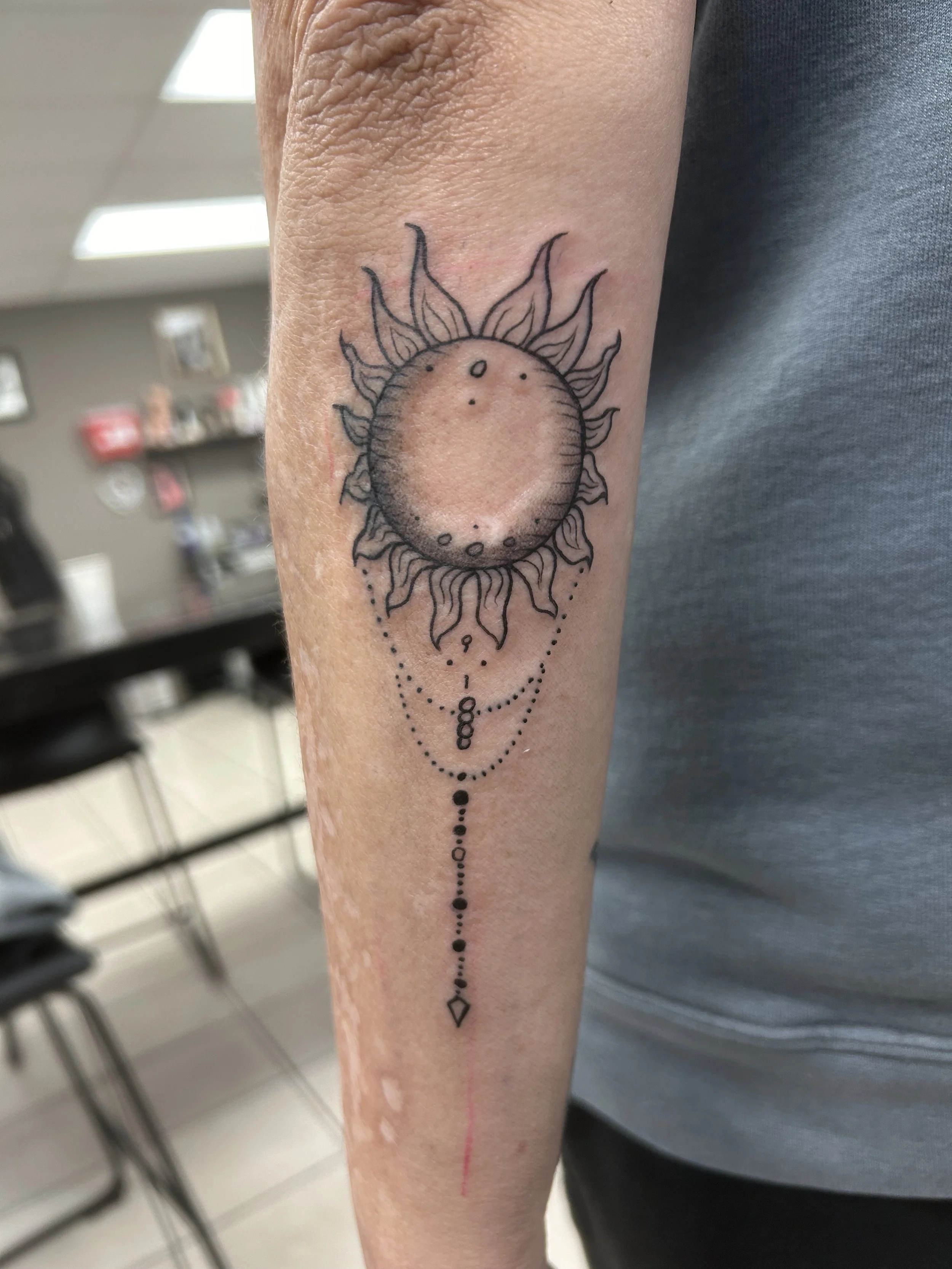 Tattoo of a sun with a crescent moon in the center, surrounded by tribal-style flames, and hanging beaded jewelry with a diamond-shaped pendant.