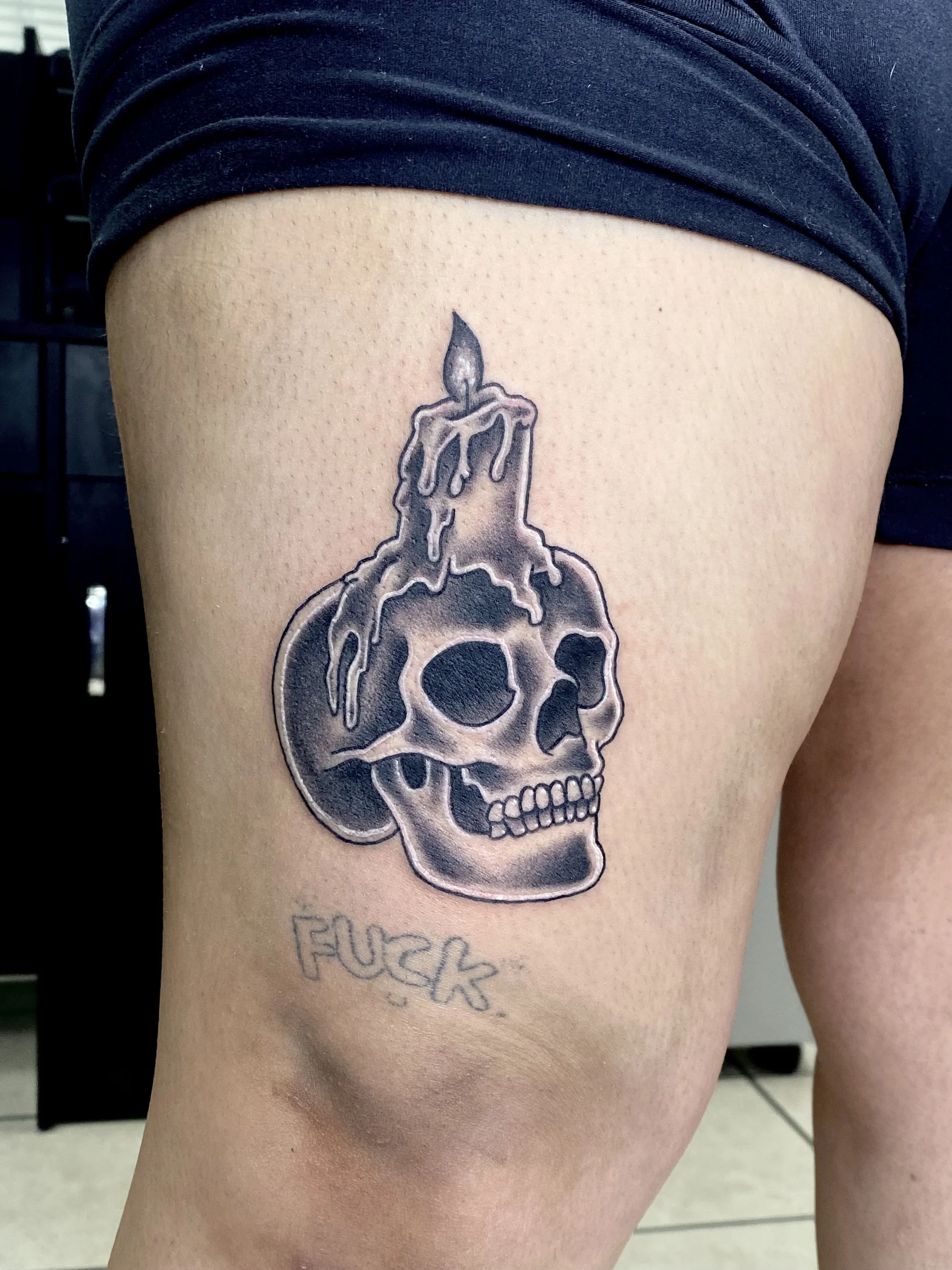 Tattoo of a skull with melted candle wax on top, with a lit candle wick, and the word 'FUCK' written underneath.