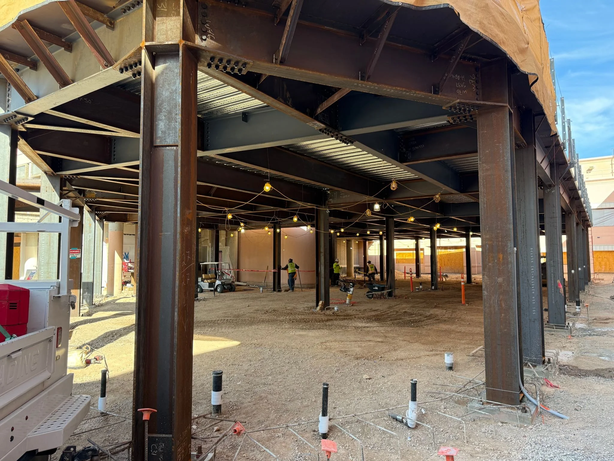 Construction site with steel framework for a building, workers in safety vests, construction equipment, and string lights inside the building.