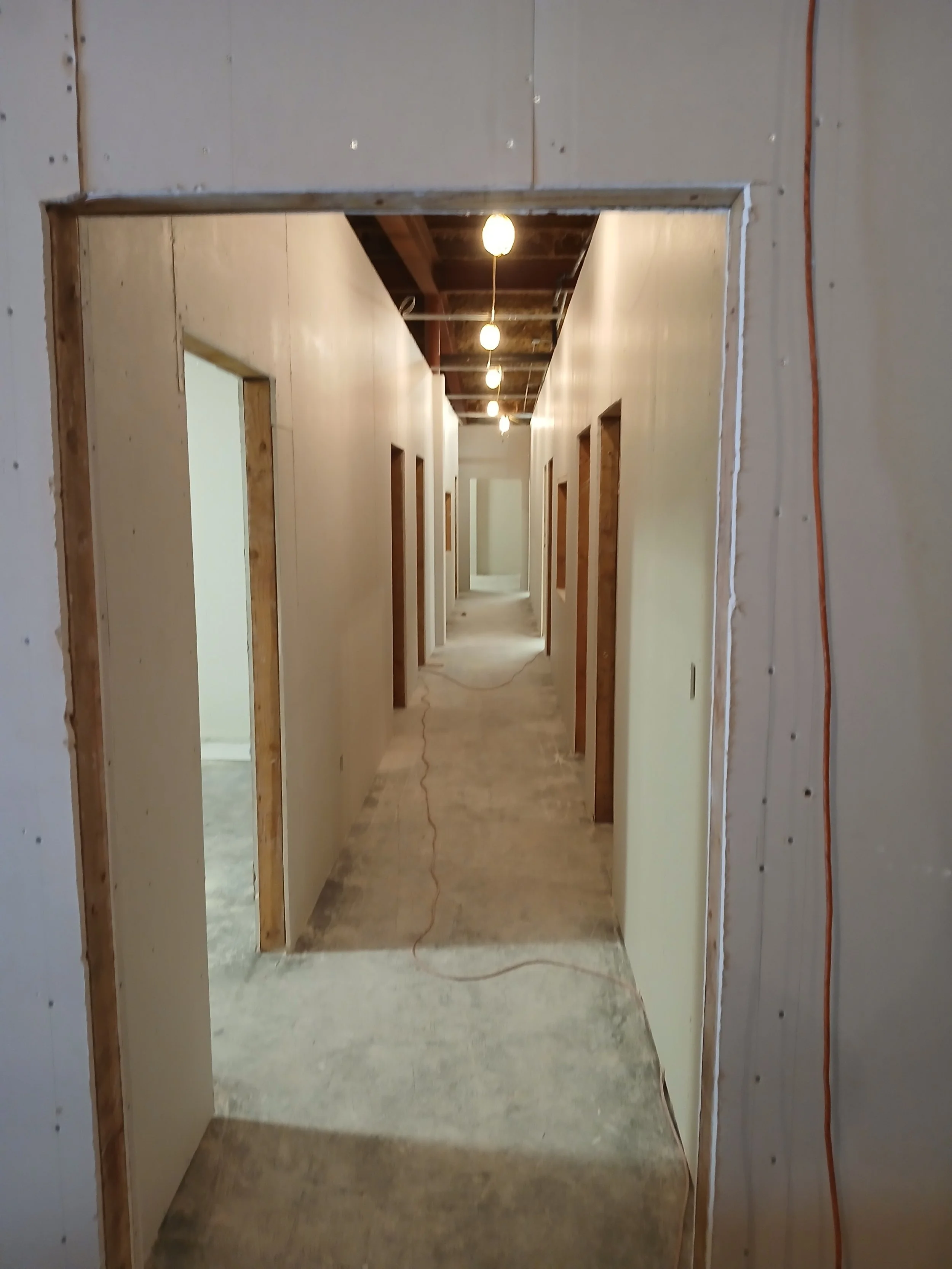 Hallway under construction with open doorways, exposed ceiling with lights, and unfinished flooring.