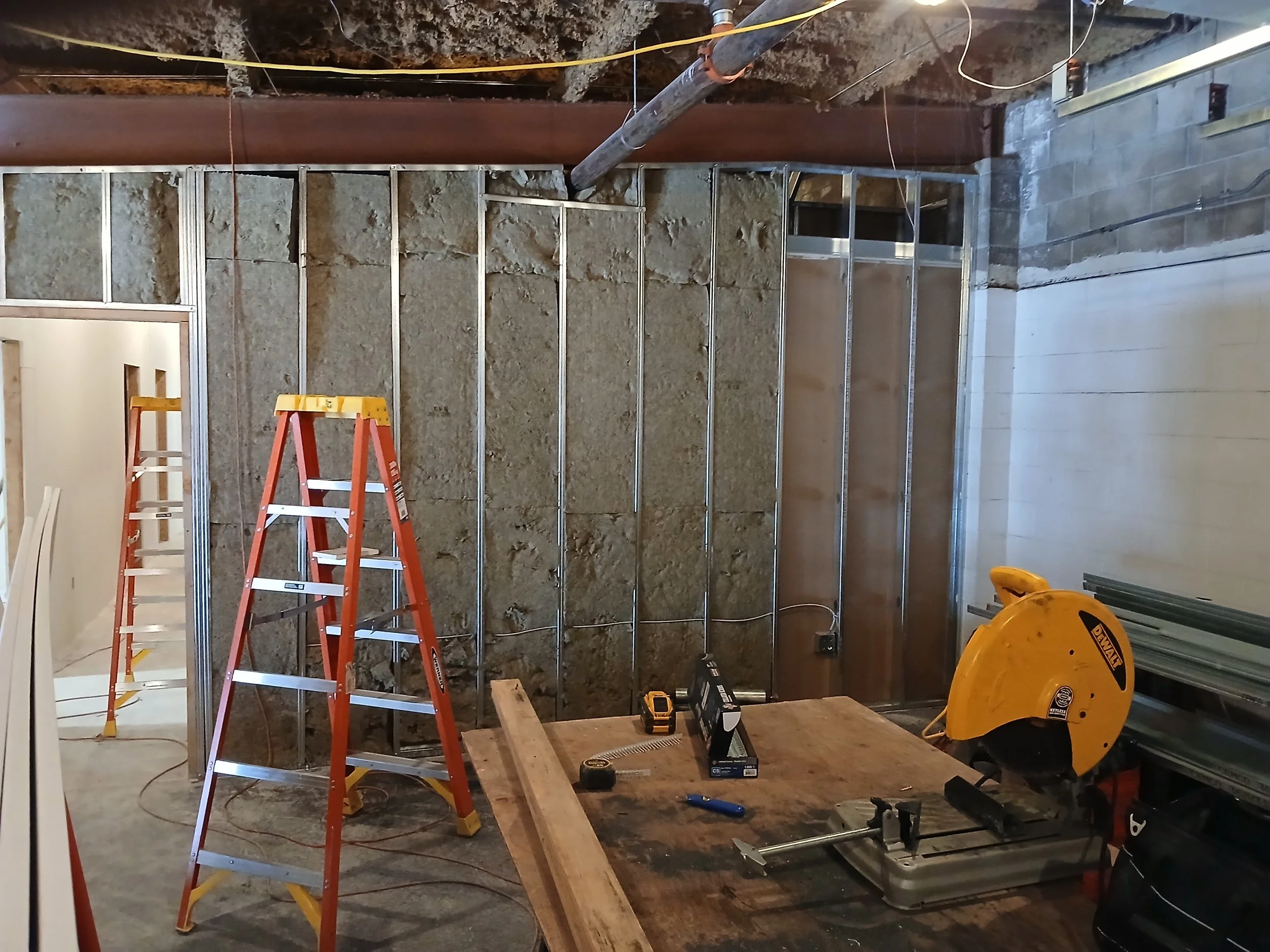 Construction site with metal wall framing, ladders, and tools, including a circular saw and measuring tape, indicating ongoing building work.