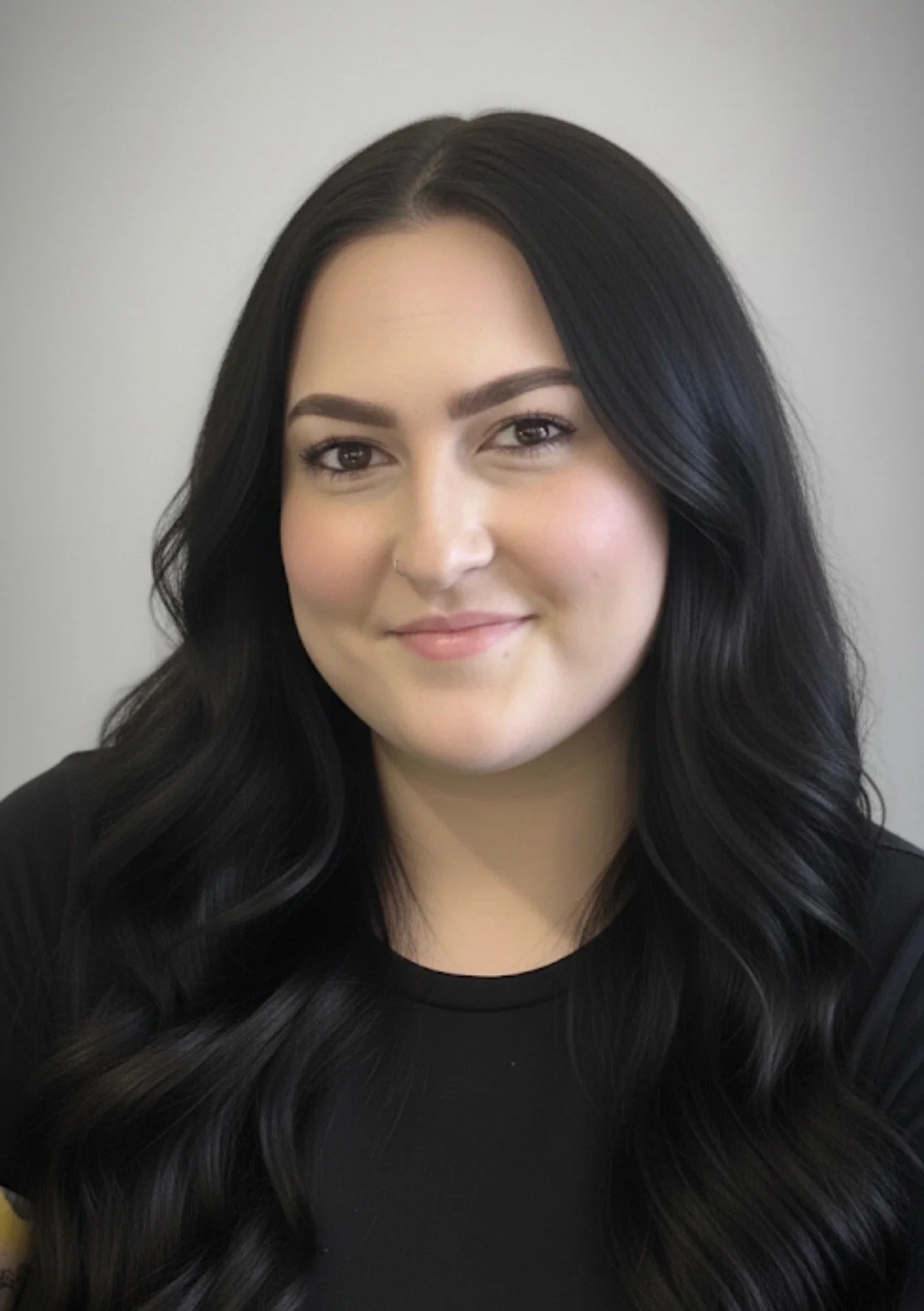 Portrait of a smiling woman with long black hair, wearing a black top, in front of a plain gray background.