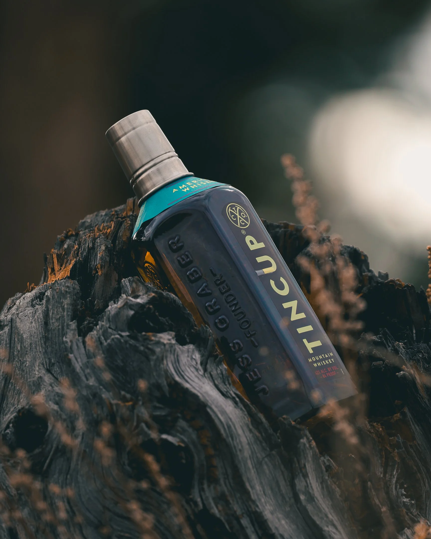 A bottle of Tincup Mountain Whiskey resting on a charred log with blurred natural background.