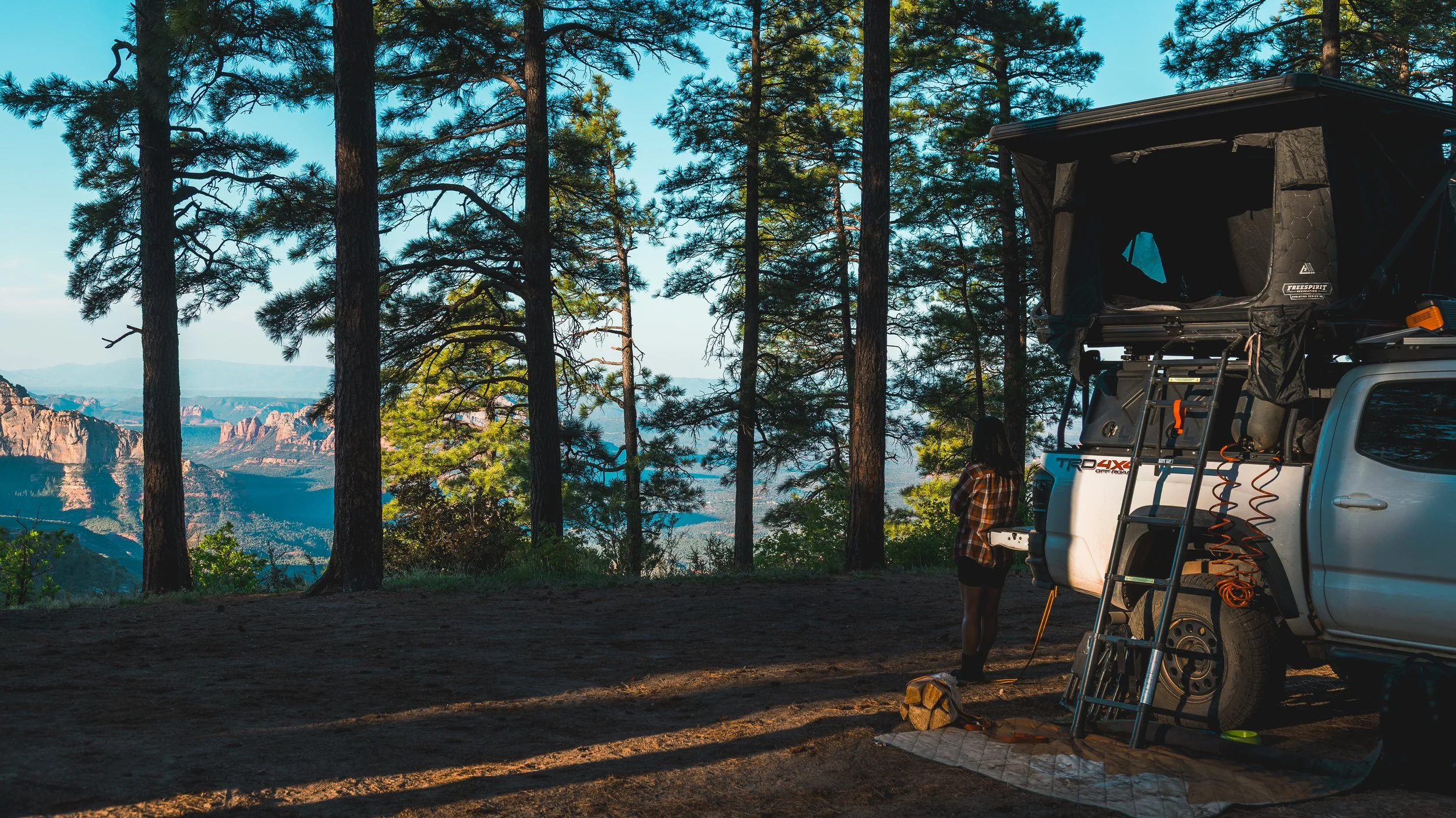 A person standing next to a pickup truck with a rooftop tent, set up in a forested area with tall pine trees and a view of mountains and a lake in the background.