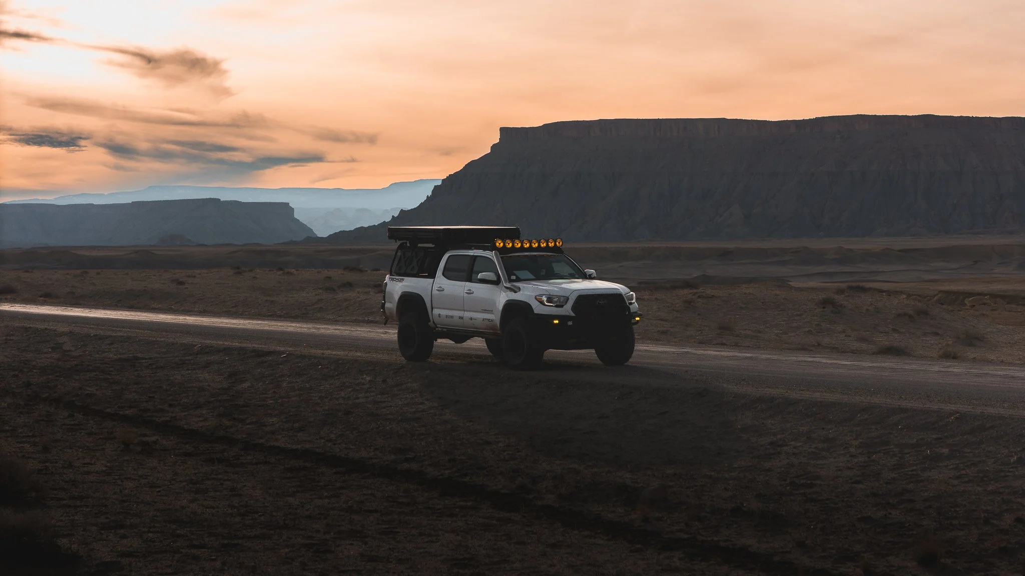 White Toyota Tacoma overlanding truck driving on a desert dirt road at sunset with dramatic canyon cliffs in the background