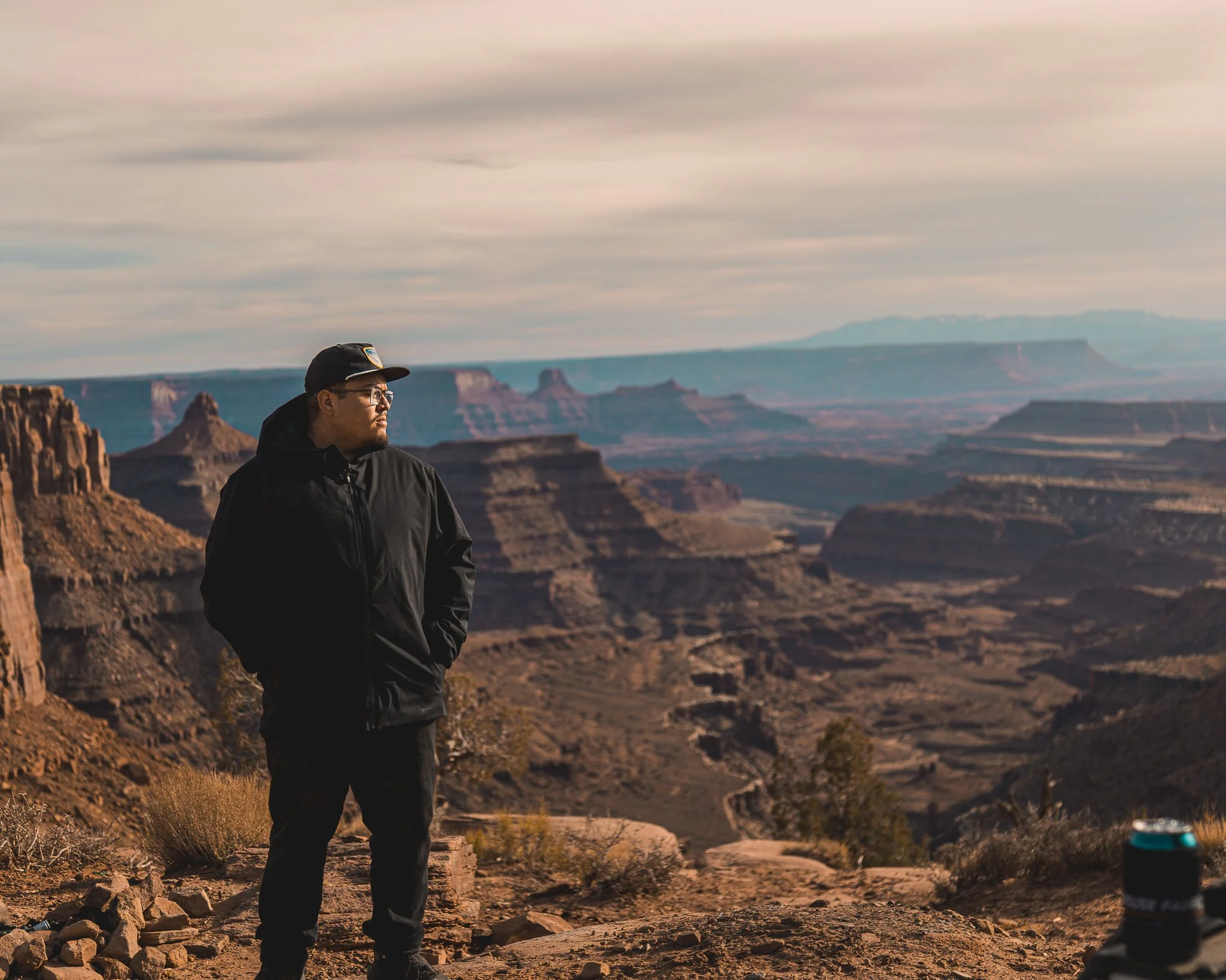 Colorado adventure photographer standing at a dramatic desert canyon overlook during golden hour with layered red rock formations in the background.
