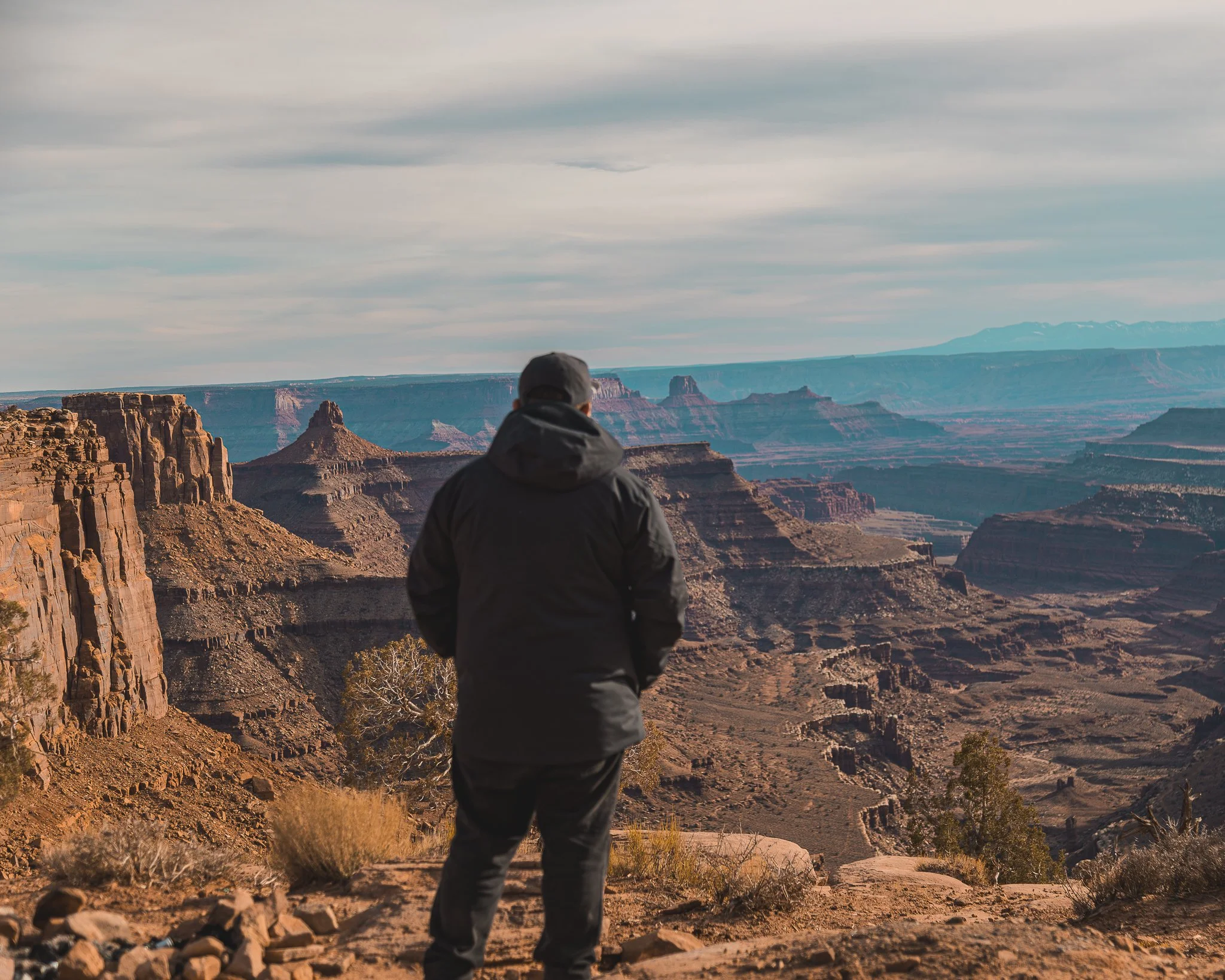 A person in a black jacket and cap standing at the edge of a canyon, looking out over the vast landscape with layered rock formations and distant mountains under a cloudy sky.