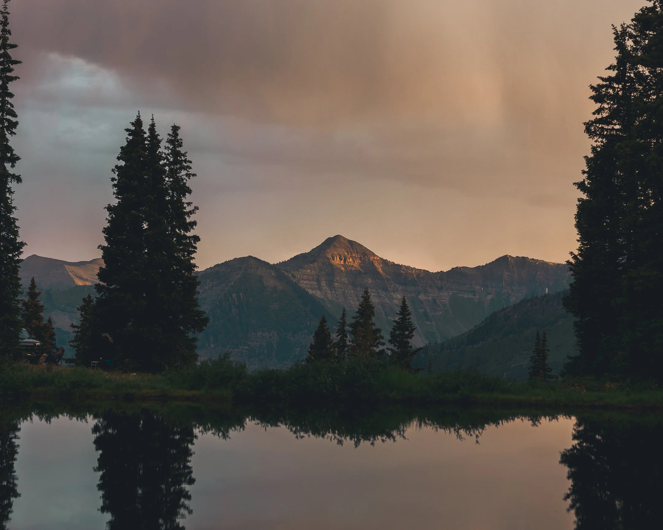 A scenic view of mountains during sunset, with trees in the foreground reflected in a calm body of water.