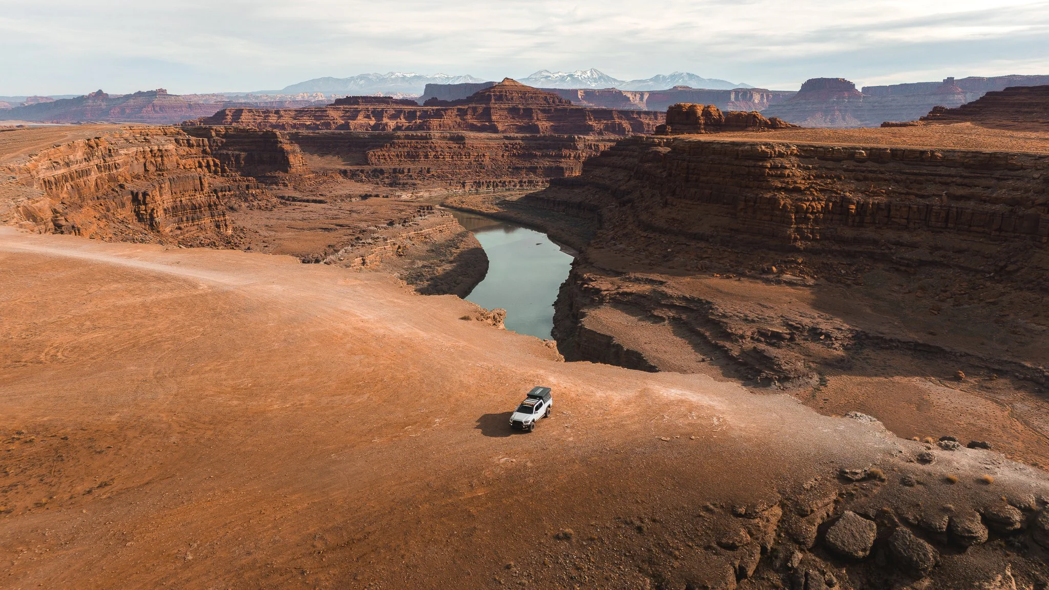 A white off-road vehicle parked on a dirt trail overlooking the Grand Canyon with the Colorado River winding through the canyon and snow-capped mountains in the background.