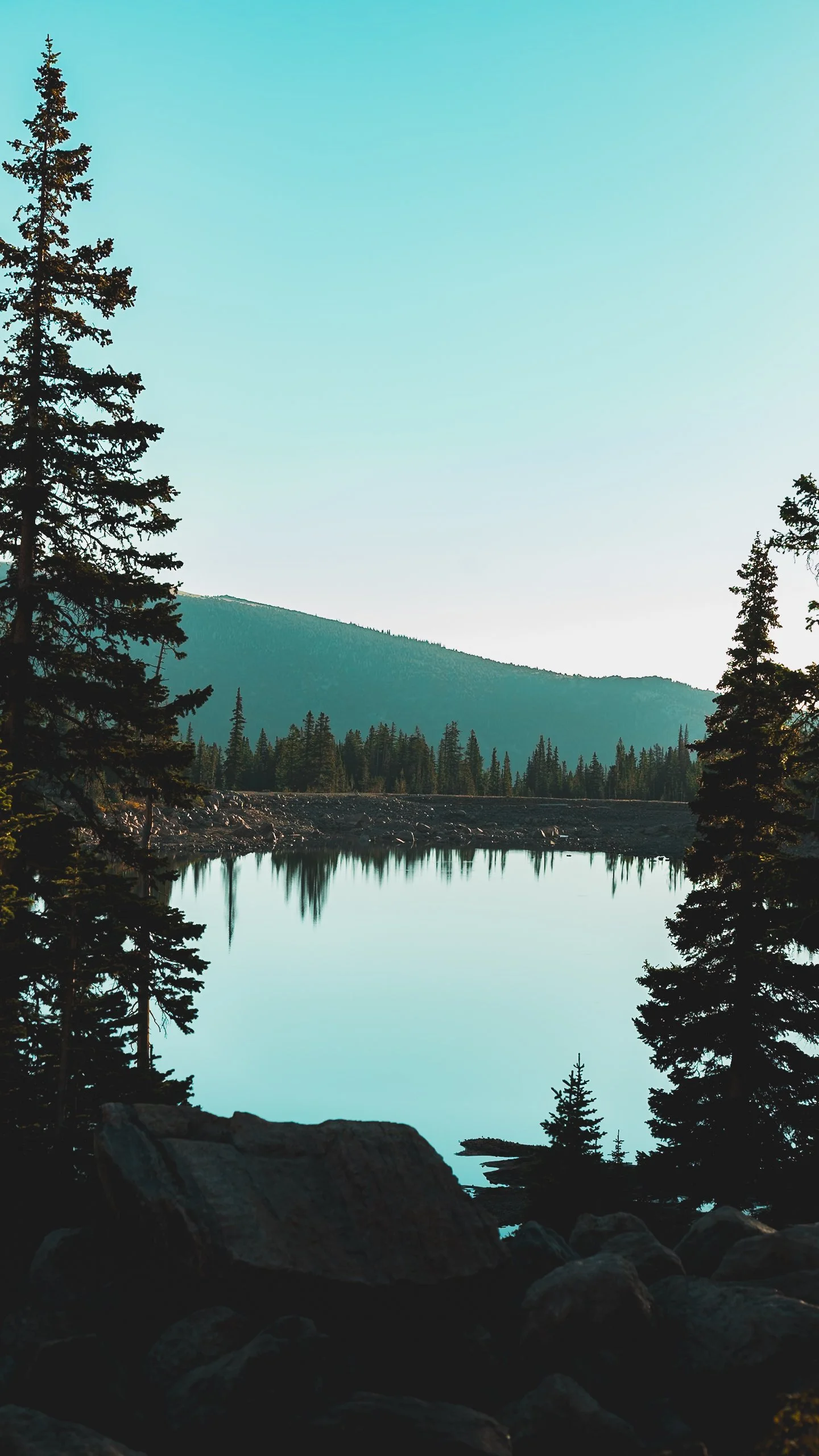 Calm mountain lake at sunrise reflecting pine trees and distant hills with rocky shoreline in the foreground.