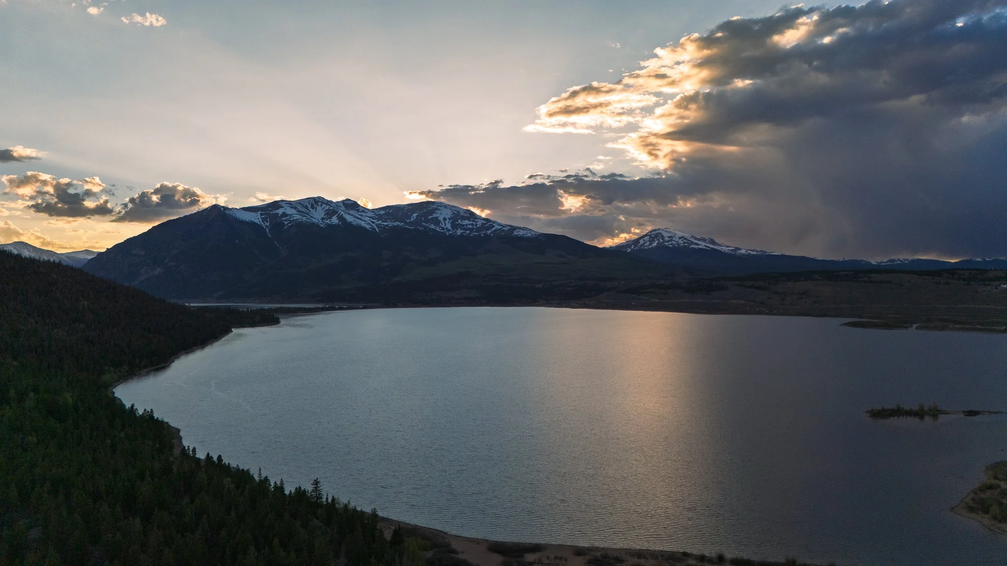 Sunset over a mountain lake with snow-capped peaks and cloud-filled sky.