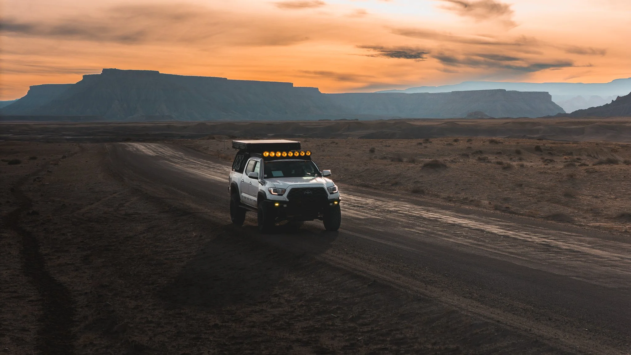 An off-road vehicle driving on a dirt trail in a desert landscape at sunset, with mountains in the background and a partly cloudy sky.