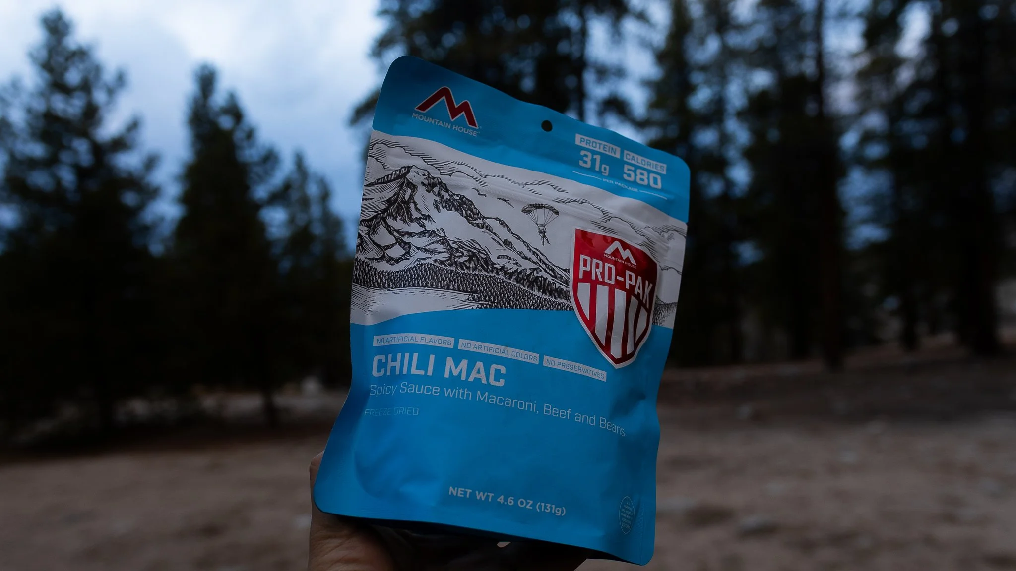 Hand holding a blue Mountain House Pro-Pak Chili Mac freeze-dried meal package with mountain and landscape illustration, outdoor environment with trees in the background.