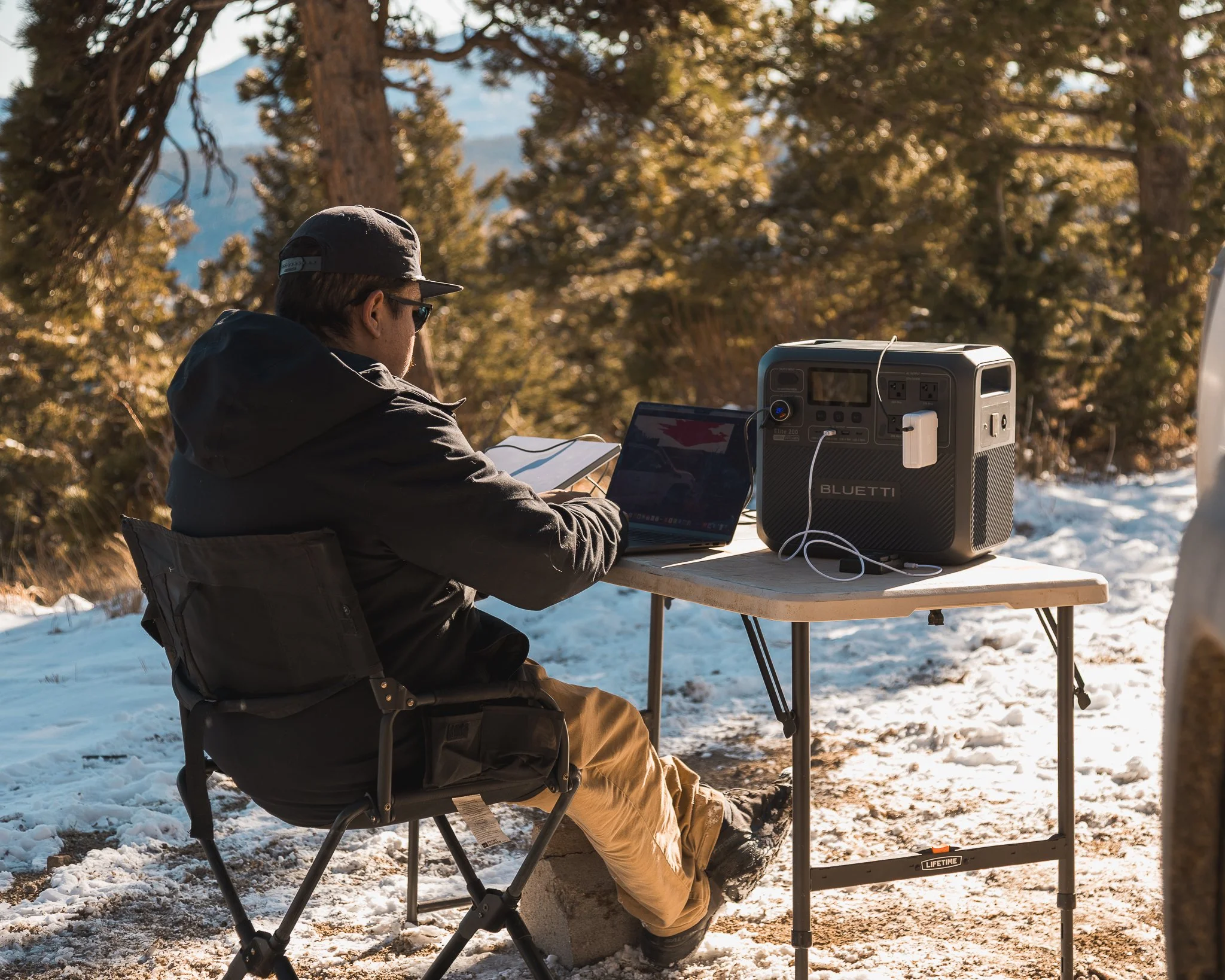 Outdoor photographer editing on a laptop in a snowy mountain forest while powering gear with a portable power station.