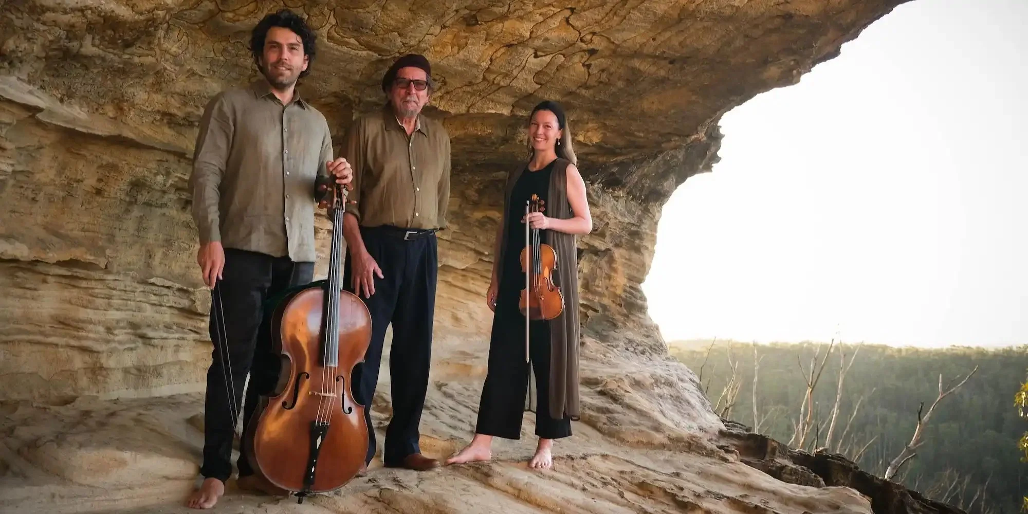 Three people, wearing neutral colours, are standing below an overhanging rock formation. One person holds a violin and another holds a cello.