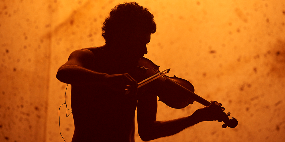 A photo of a person playing the violin, silhouetted by a background lit with warm orange light.