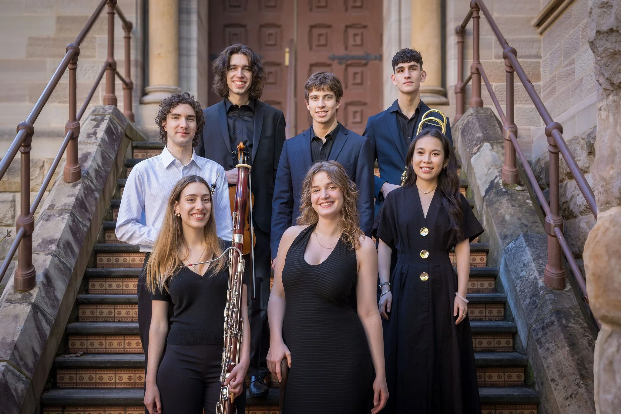 A group of musicians are arranged on a stairway leading to the entrance of a historic building. They smile directy at the camera, holding their instruments by their sides.