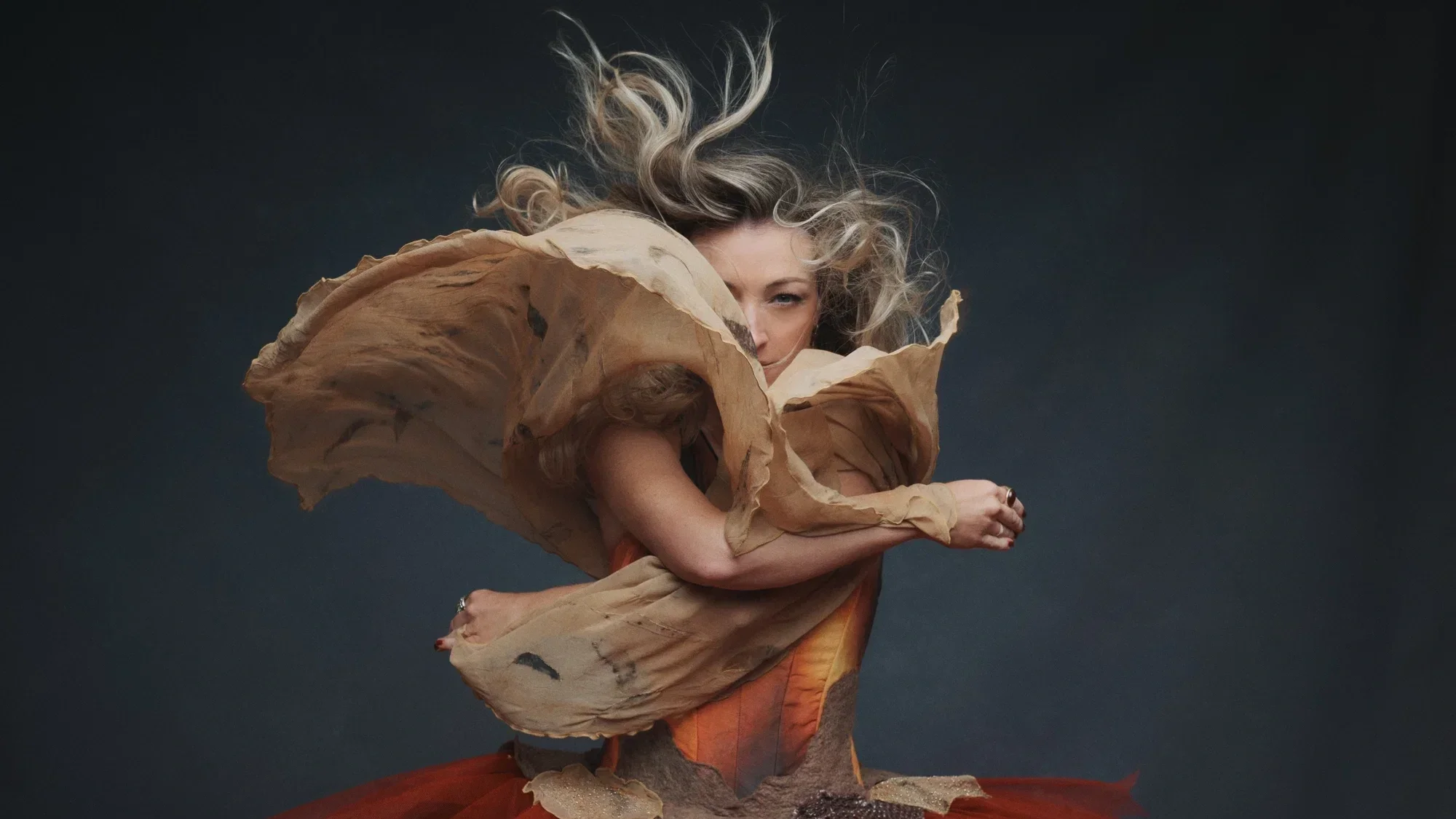 A dramatic photo of Kate Miller-Heidke. Her face is partially covered by a piece of fabric as she twirls in a layered costume of earthy colours.