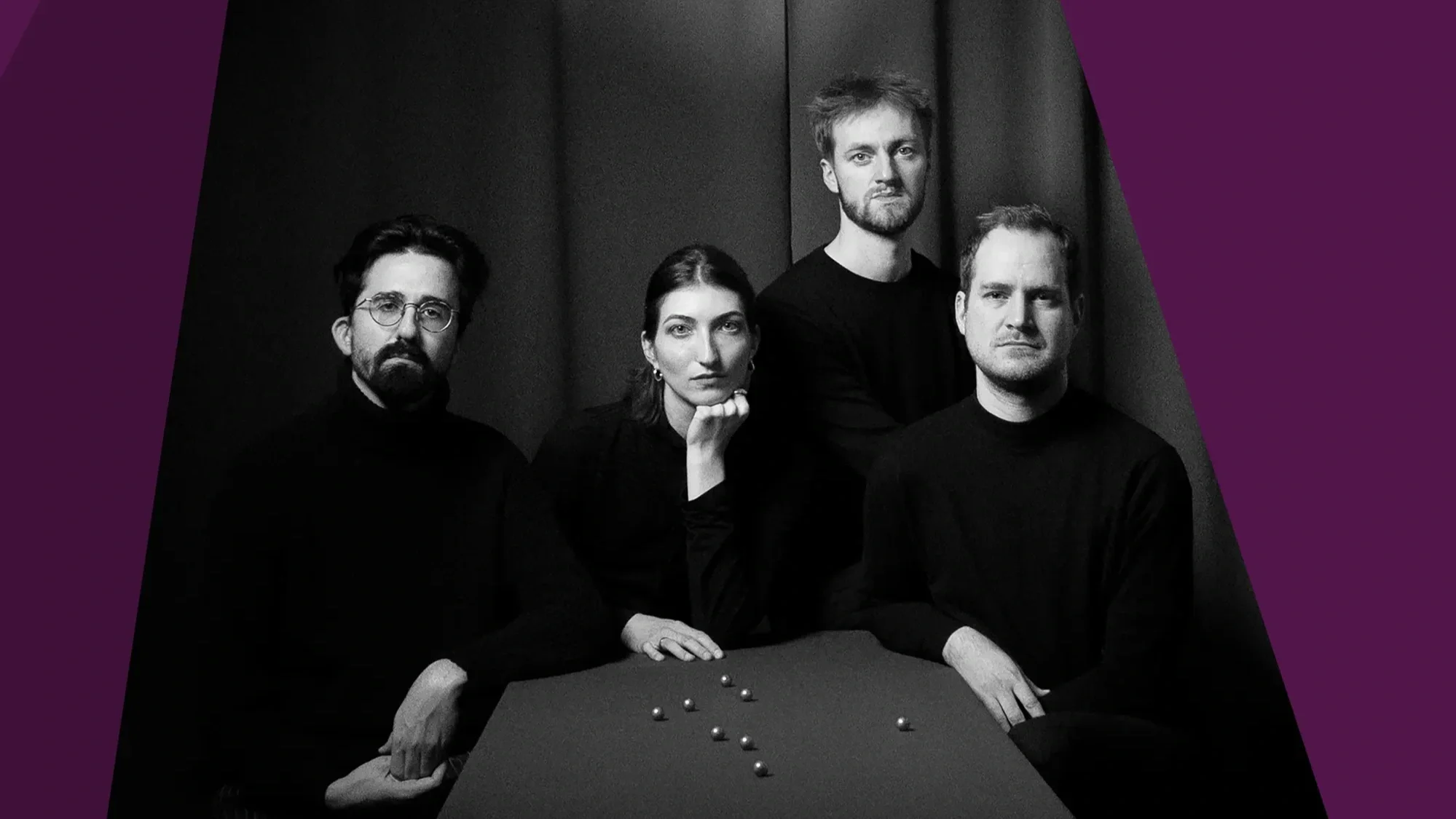 A black and white photograph of a group of four people wearing plain black clothing. They are seated by a table that displays a collection of marbles. Purple graphic panels frame the image.