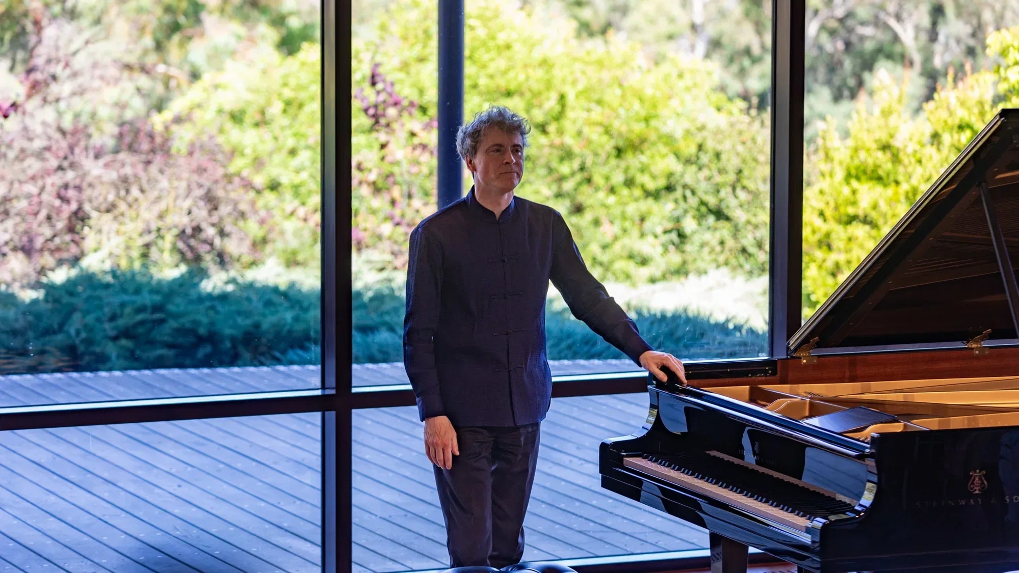 A photo of Paul Lewis standing next to a grand piano. Large glass windows reveal an outdoor garden setting.