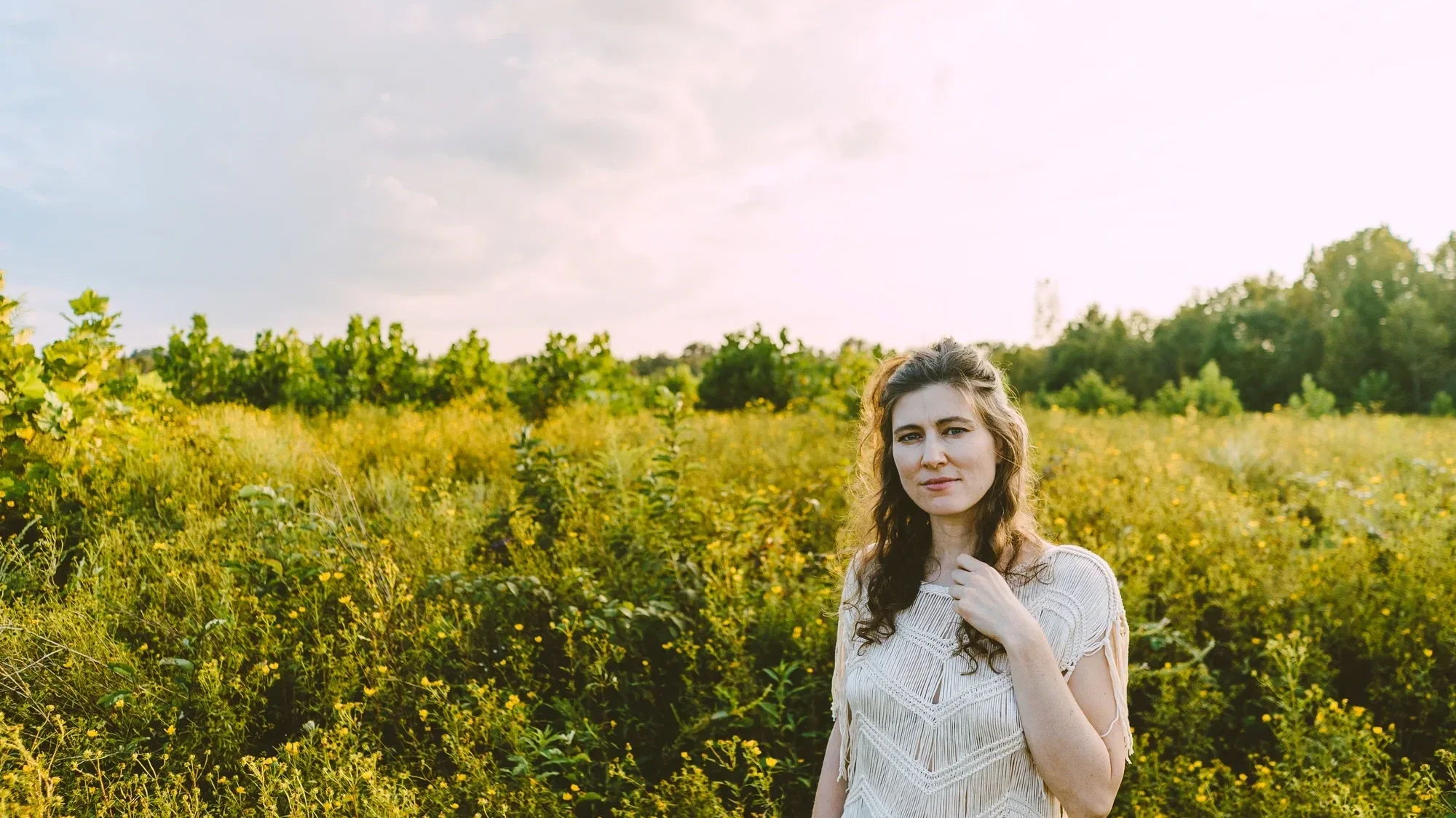 A photo of Ruth Moody wearing a fringed top, standing in a field of yellow flowers. The scene is softly lit with golden light.