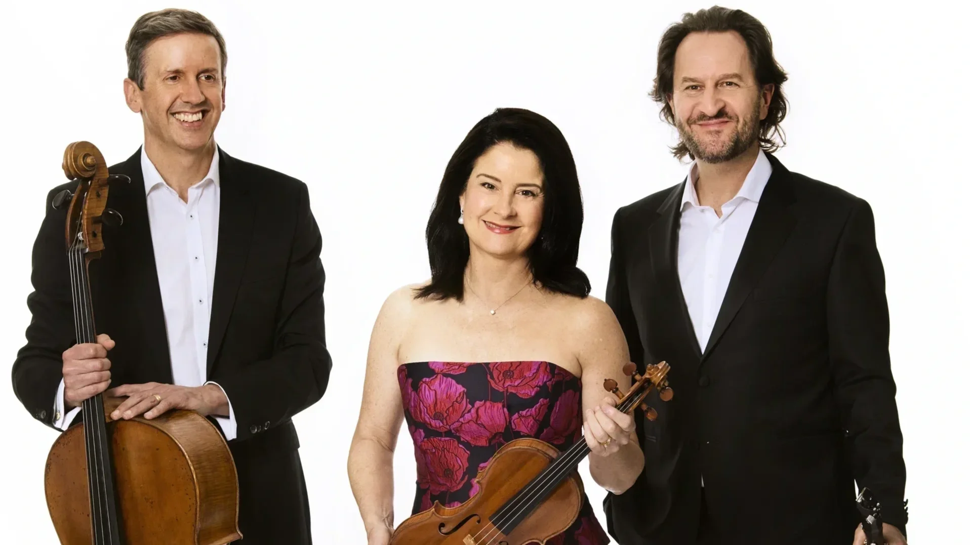 A trio of musicians stand side-by-side, holding their instruments in front of them. They are dressed formally, standing against the white background.