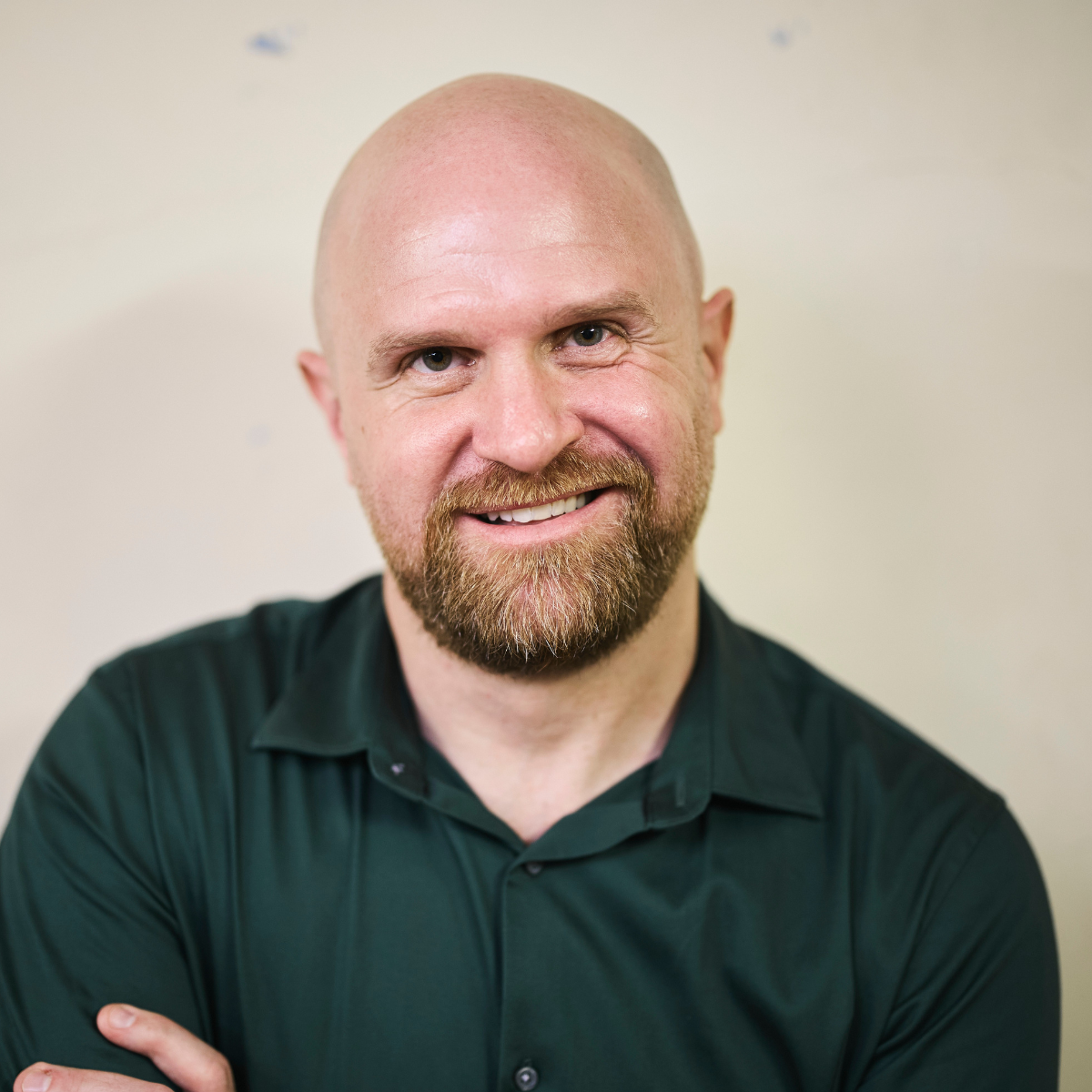 A bearded man wearing a dark green open collared shirt stands against a light background, smiling directly at the camera.