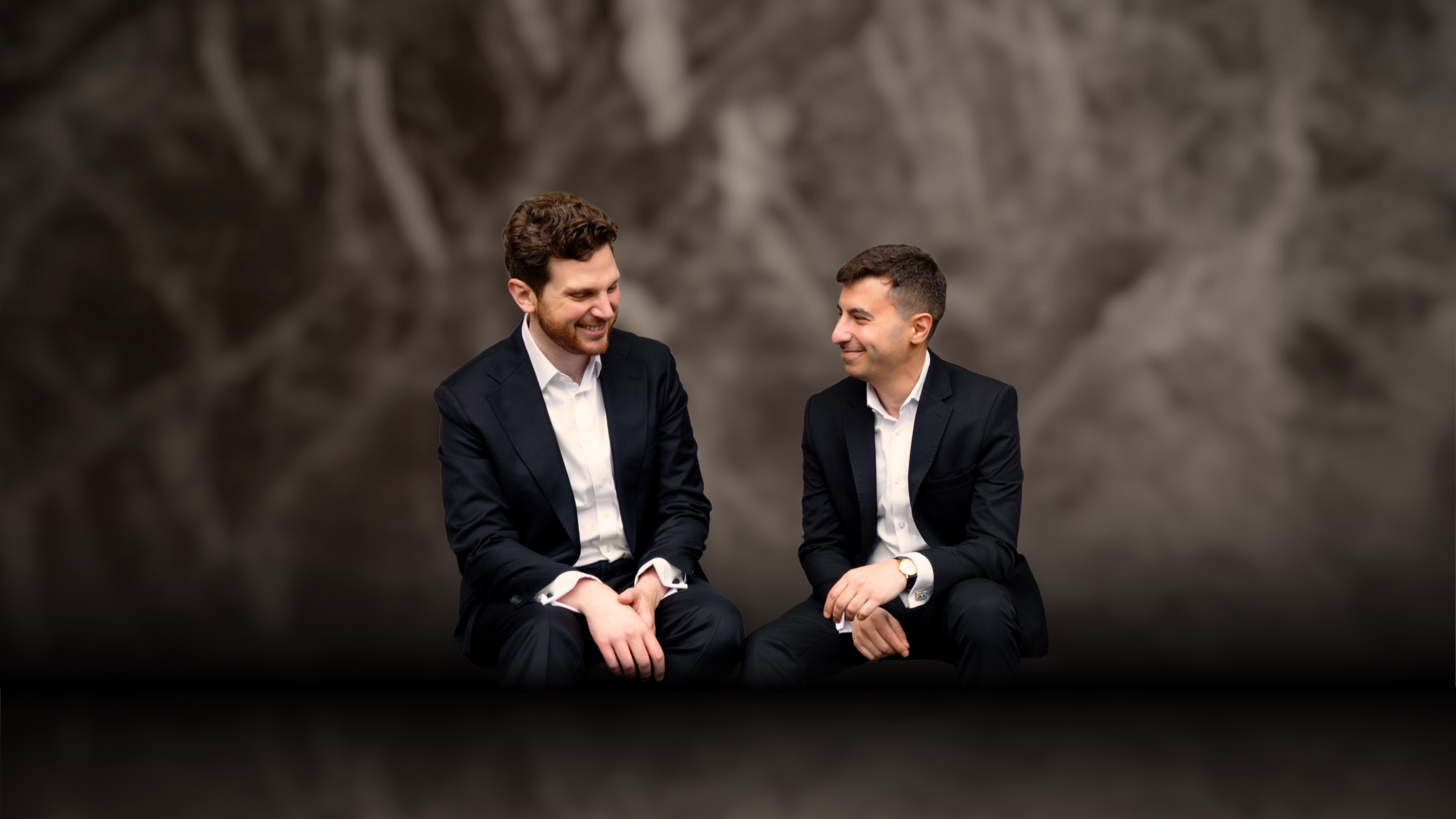 Two people wearing dark-coloured formal suits and open-collared white shirts laugh between themselves. The photo is blended with a textured grey background.