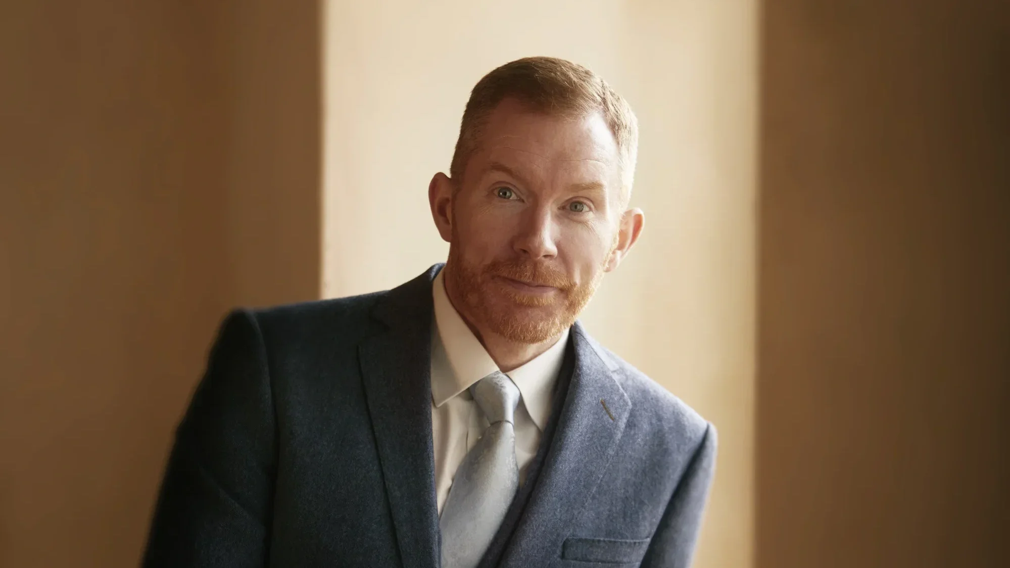 A man with red hair and and a groomed beard is wearing a blue suit and tie. There are rendered walls and shadows in the background.