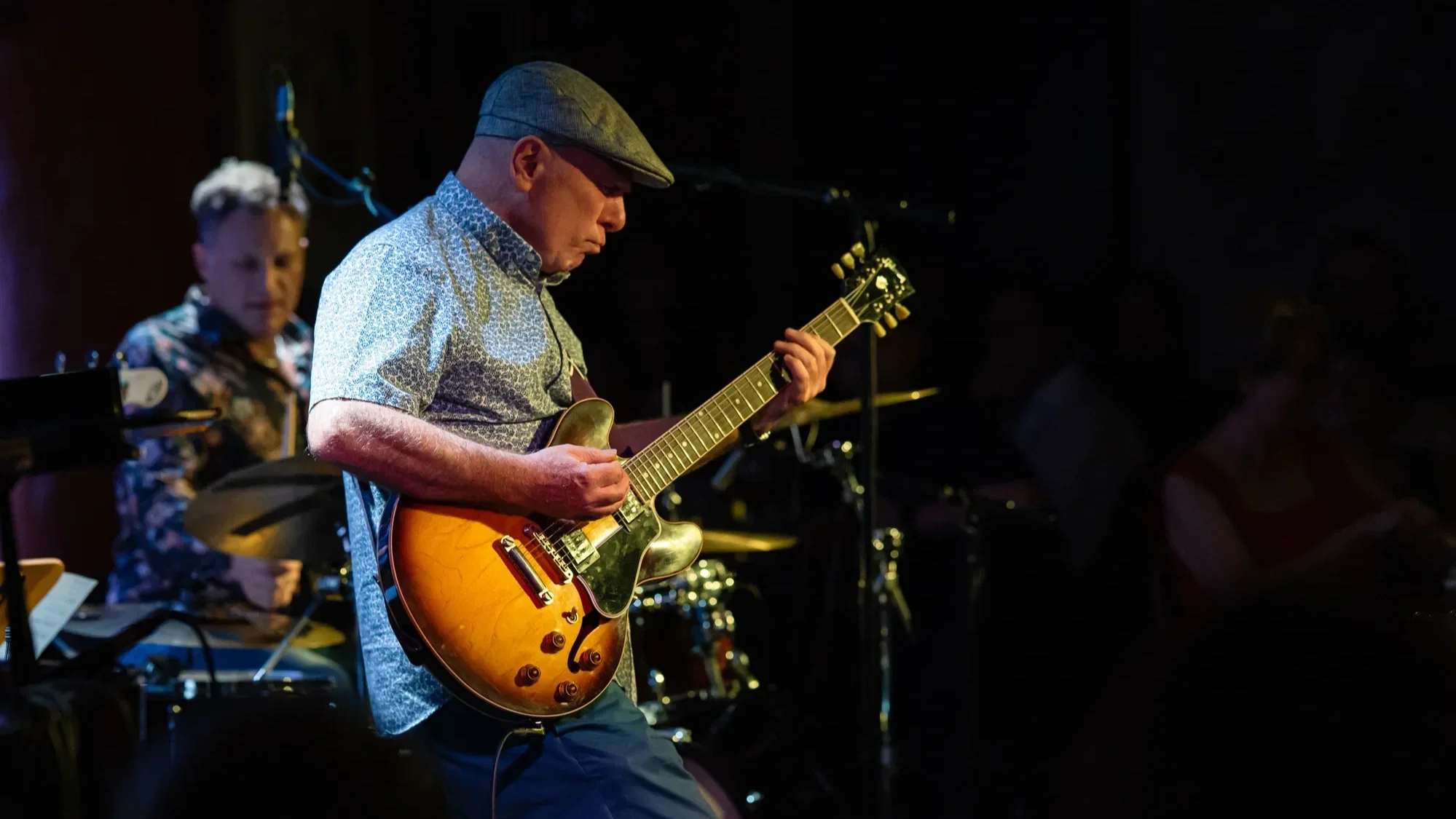 A man in a patterned, short-sleeve shirt and dark cap plays an electrical guitar. A drummer is visible in the background. Stage lighting suggests a live performance.