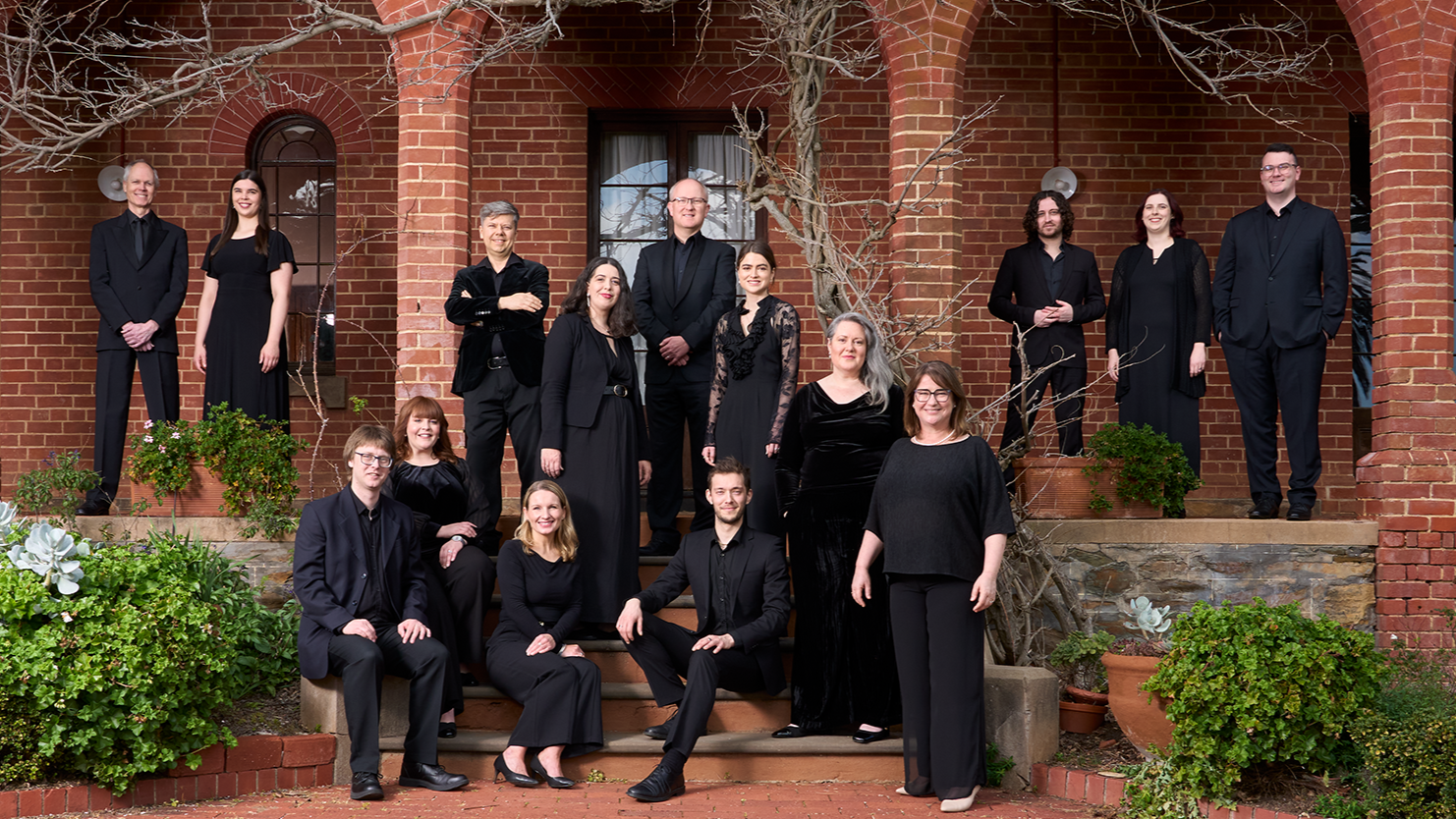 A group of people are are formally dressed in black, posing on stairs and in archways of an historic brick building.