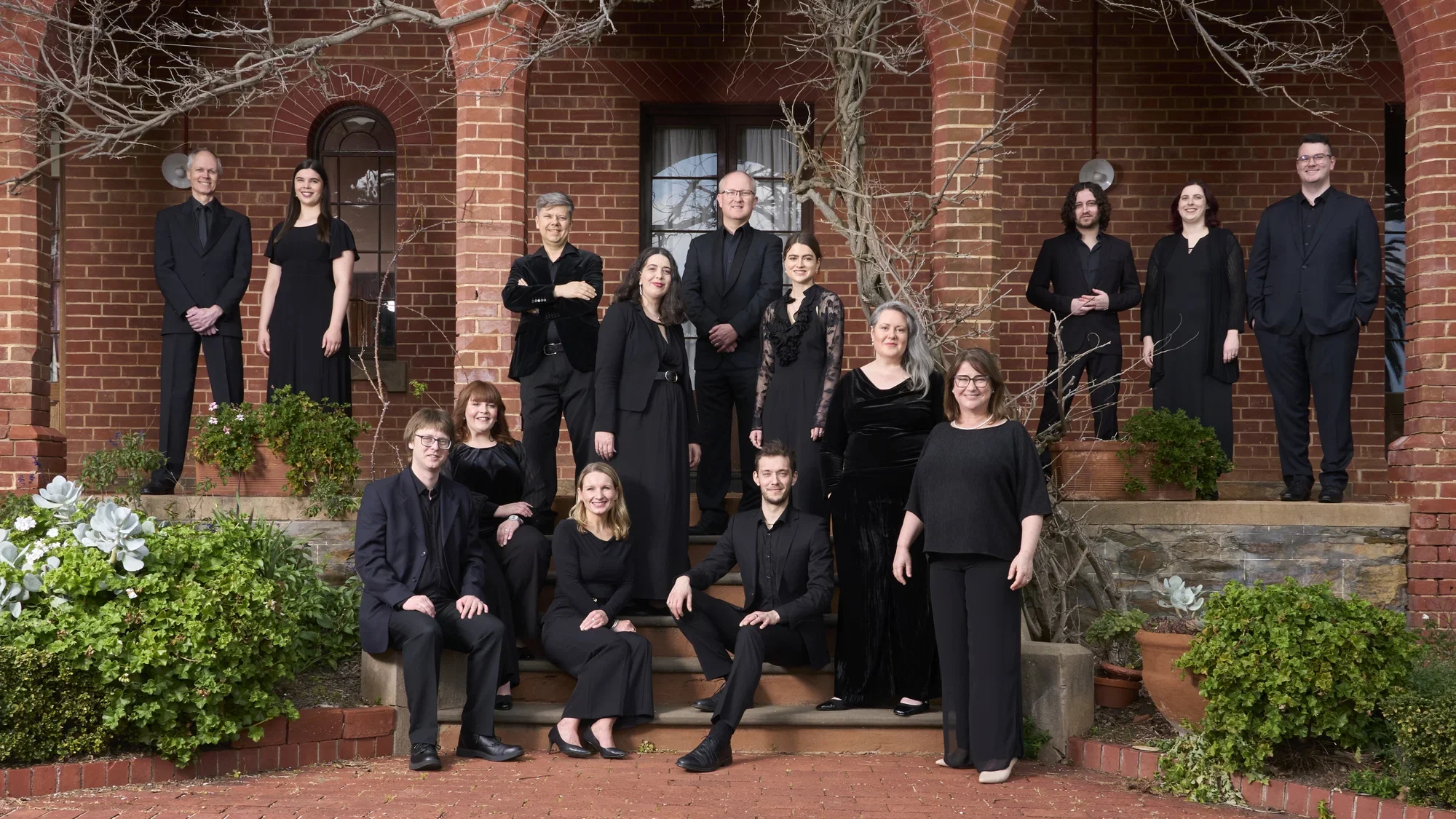 A group of people are are formally dressed in black, posing on stairs and in archways of an historic brick building.