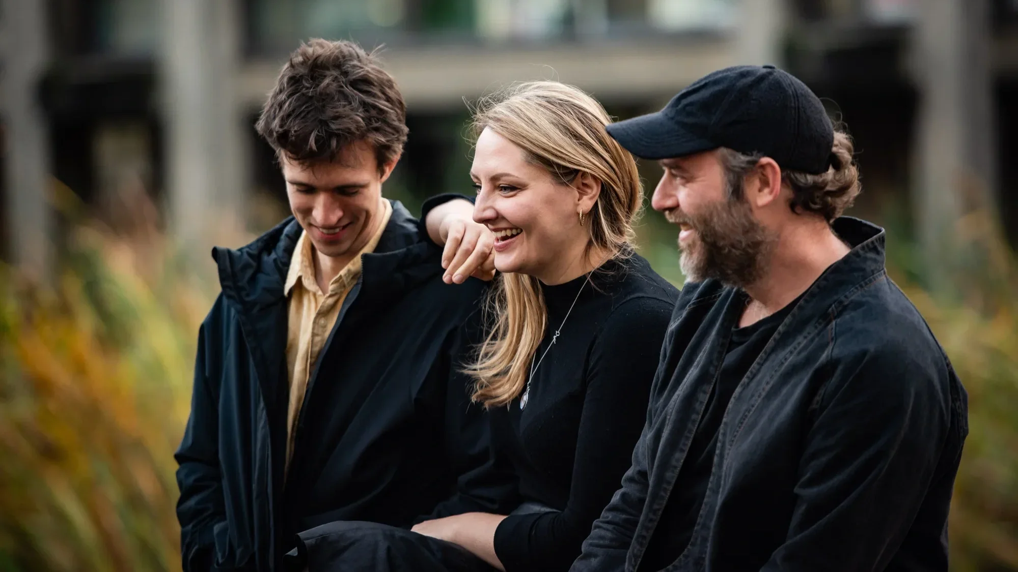 Three people with friendly smiles gather outdoors. They are wearing layers of darkly coloured clothing.