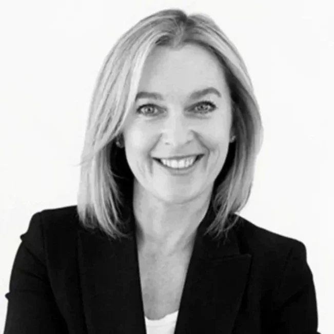 A black and white headshot of a person with shoulder length light-coloured hair smiling directly at the camera. They wear a formal dark jacket.