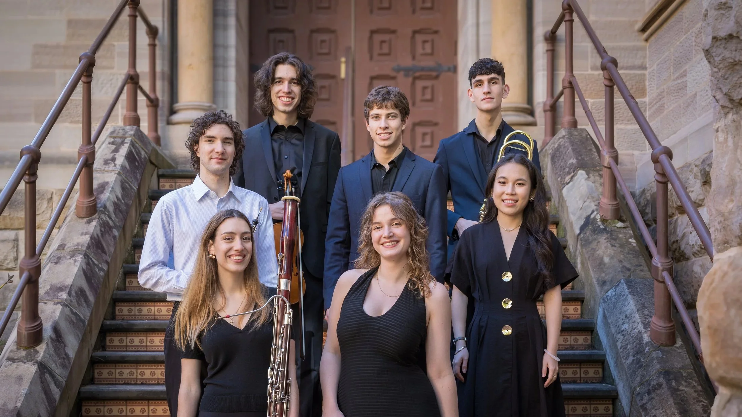 A group of young musicians are arranged on the staircase of a historic, ornate building. The individuals are dressed formally with some holding instruments.