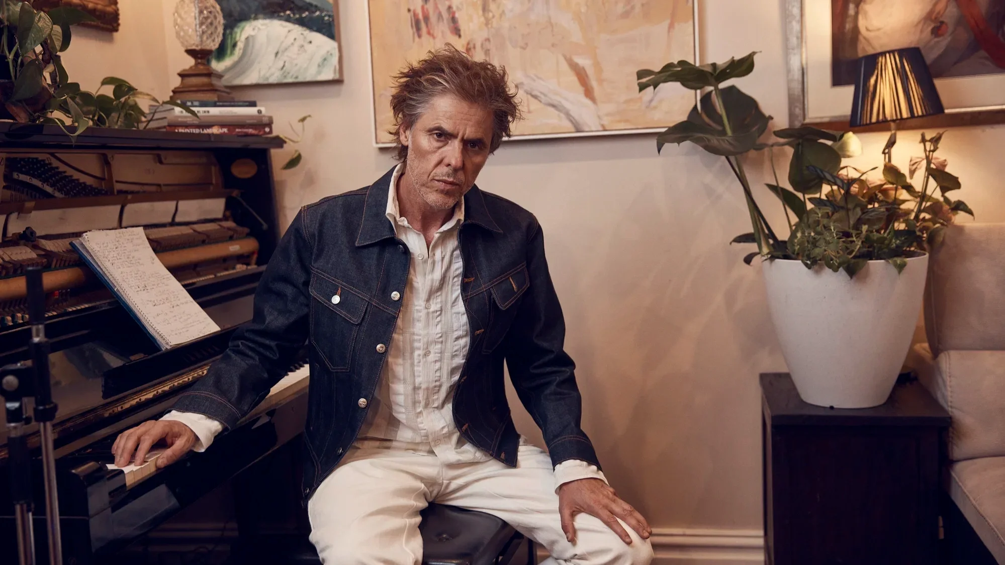 A photo of Tim Freedman seated by an upright piano. The wall behind displays several artworks and the space is decorated with lamps and potplants.