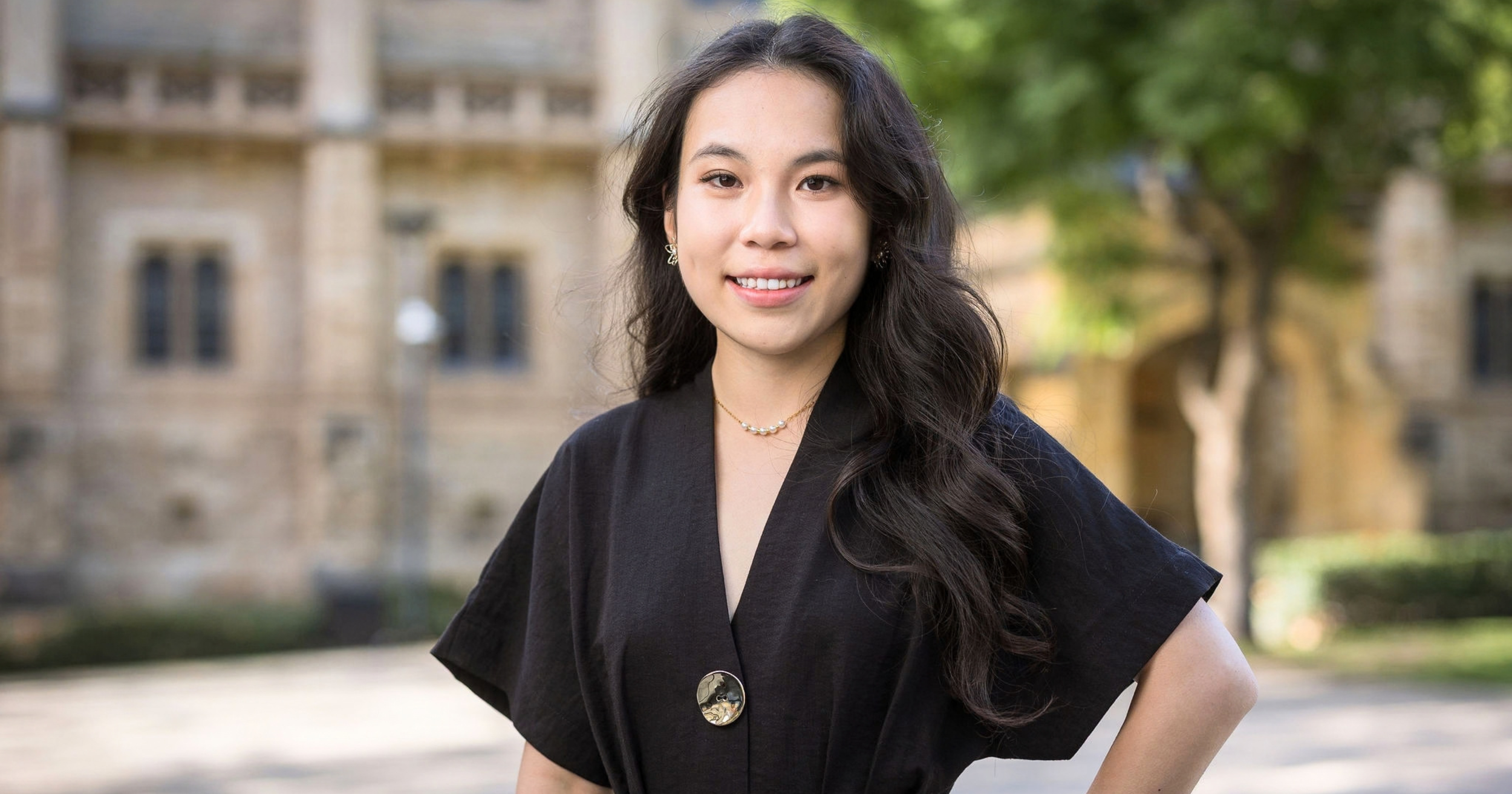 Soprano Teresa Chen is photographed wearing a black top and gold jewelery. A historic stone building is visible in the background of this photo taken outdoors by Photos by Jamois.