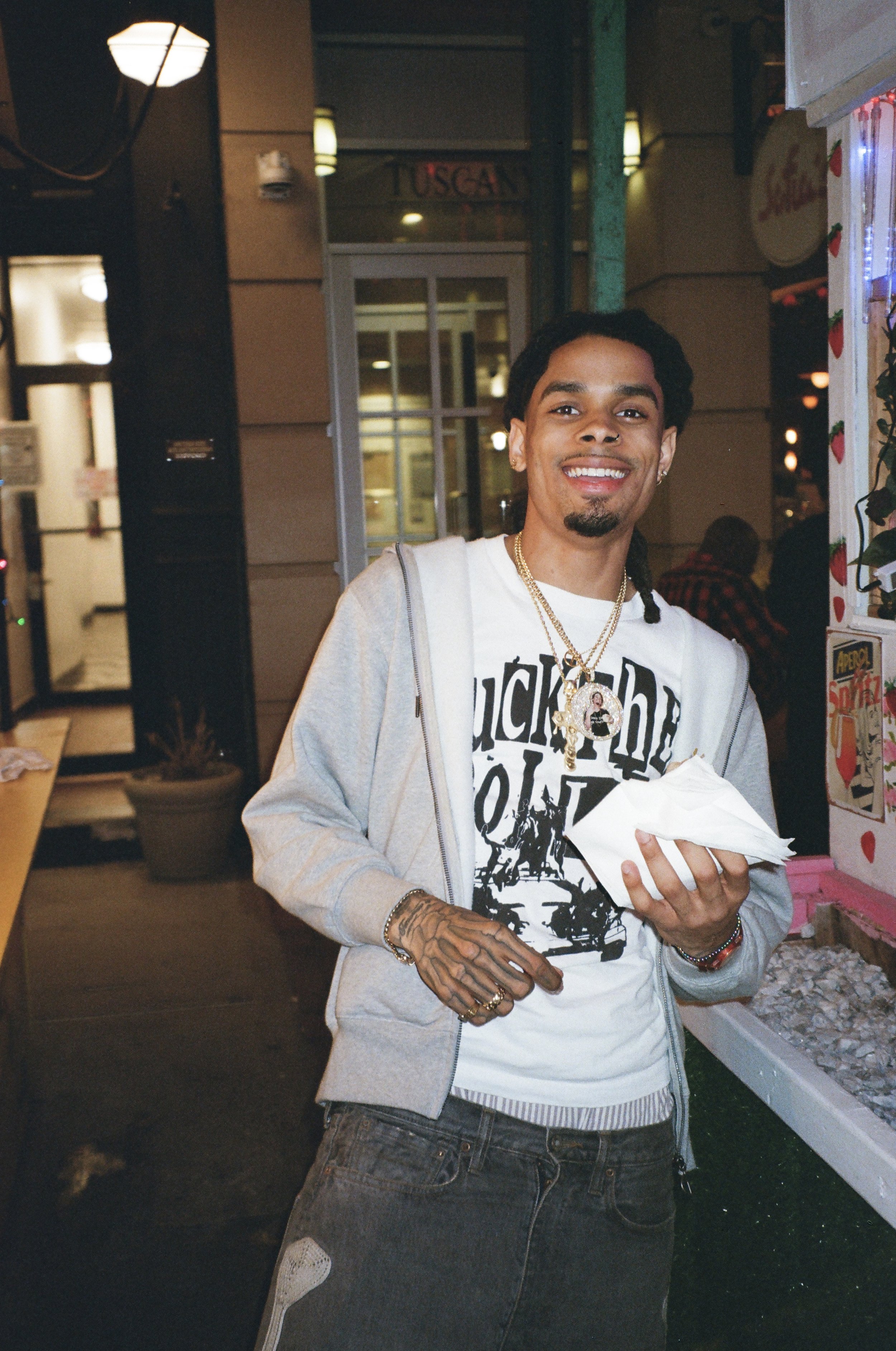 A young man smiling and holding napkins outside a restaurant at night, wearing a graphic T-shirt, gray hoodie, and necklace.