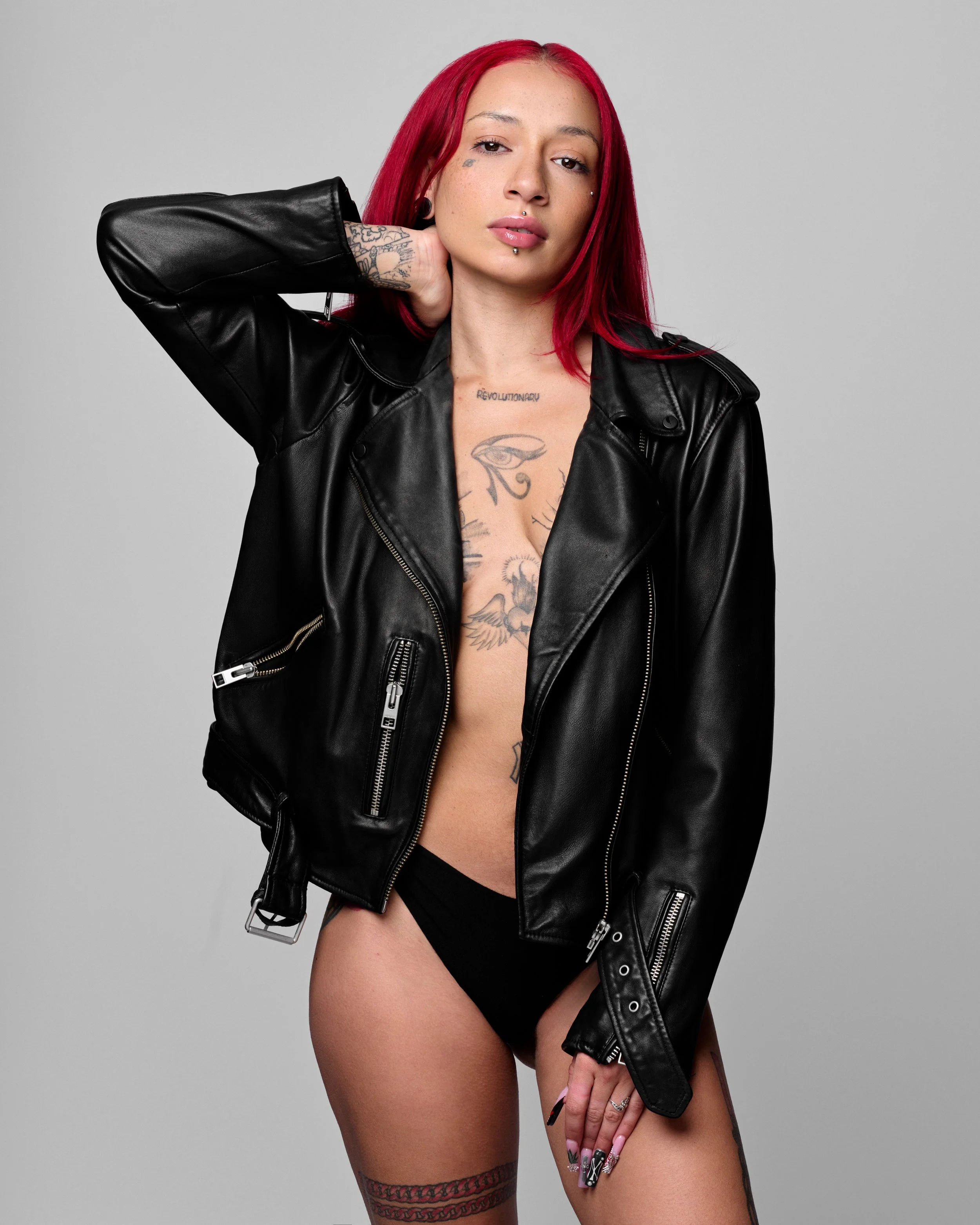 A woman with red hair and multiple tattoos wearing a black leather jacket and black underwear, posing against a plain gray background.