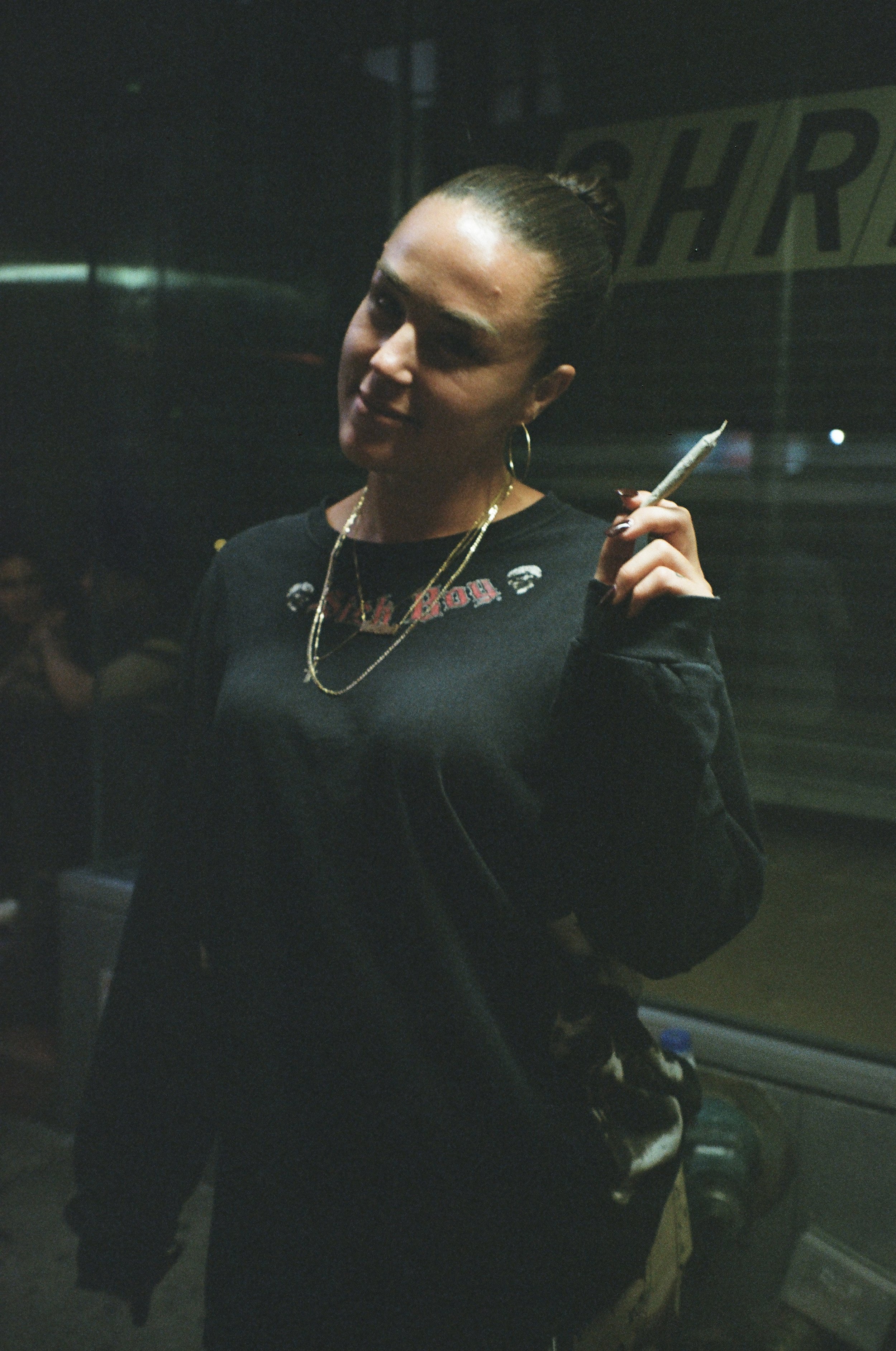 A woman with dark hair in a bun, wearing gold hoop earrings and a black sweatshirt with red text, is standing and holding a cigarette in her right hand inside a dimly lit setting. There is a partially visible sign behind her.